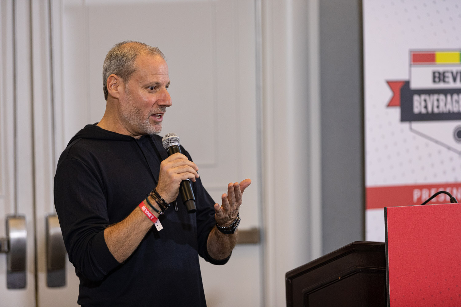 A man in a black hooded sweatshirt with a wristband holds a microphone and gestures in front of a Beverage School banner.