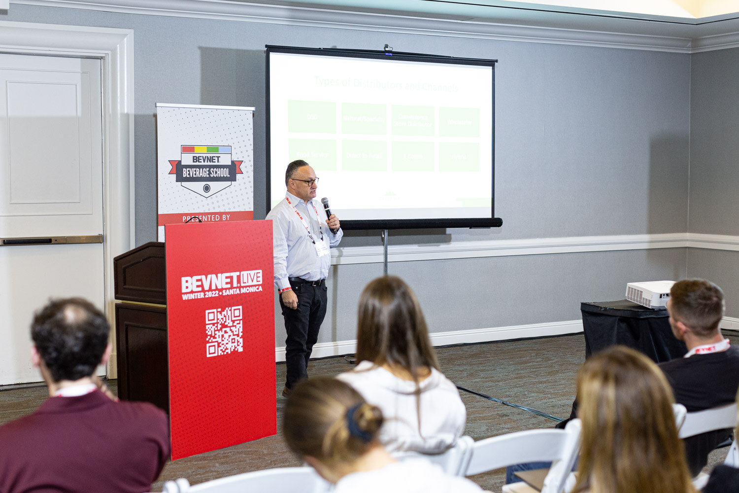 A man in a light checked shirt stands by a green slide on a projection screen, with a red BevNET Live podium and attendees in the foreground.