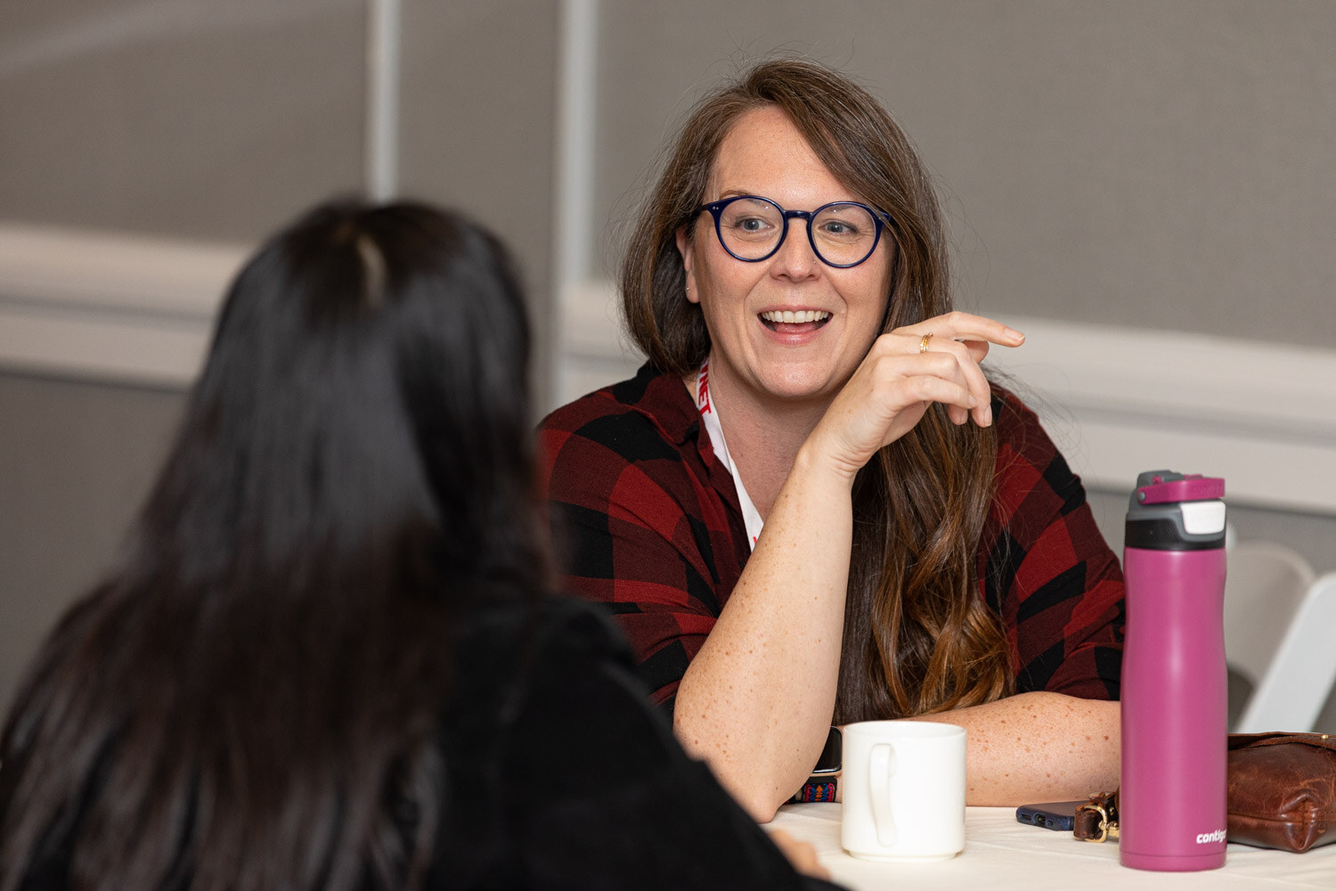 A smiling woman in glasses and a red plaid shirt sits across a table from an attendee seen from behind, with a pink water bottle and a coffee mug on the table.