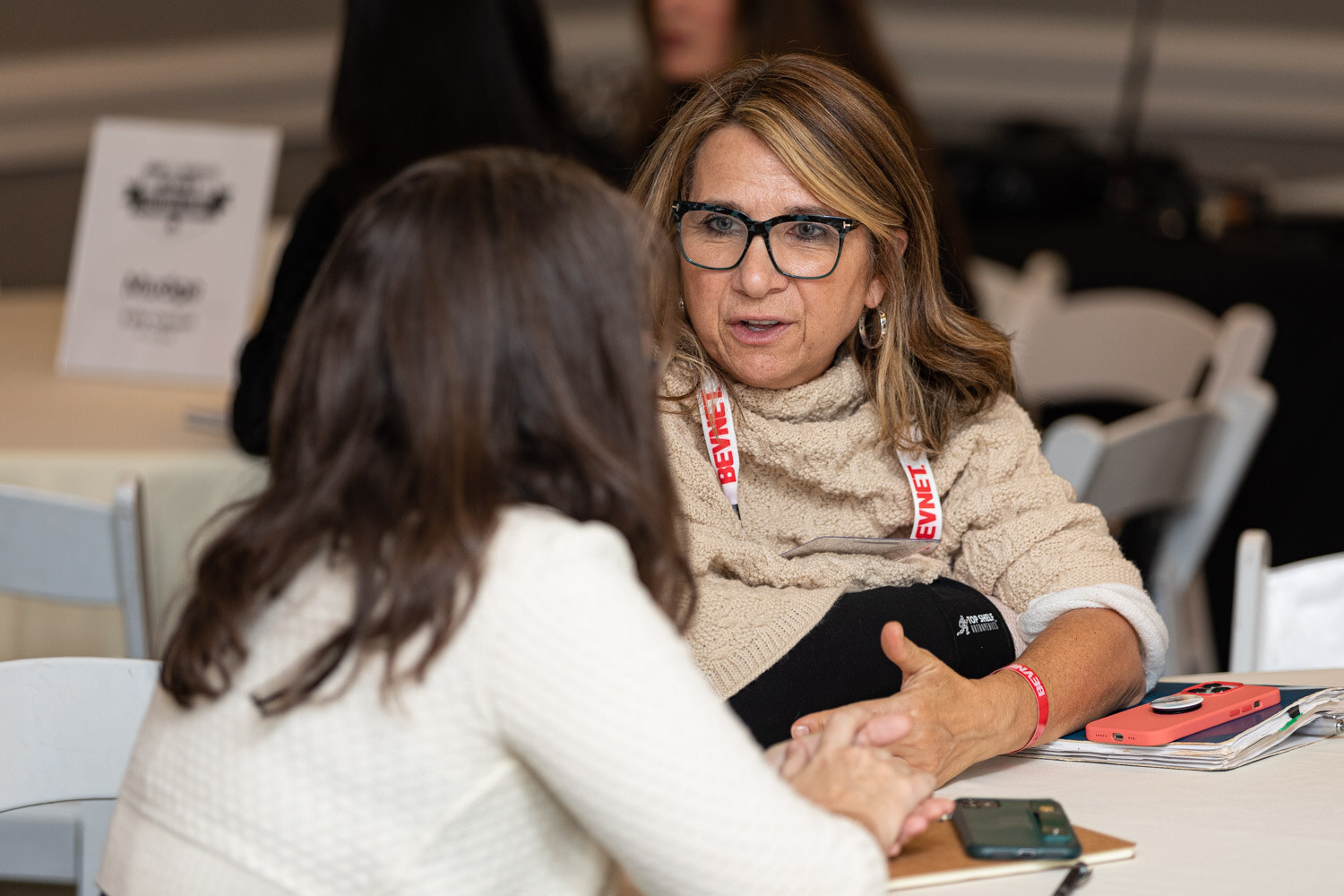 A woman in glasses and a cream sweater with a BevNET lanyard clasps hands with a woman seen from behind across a table in a meeting room.
