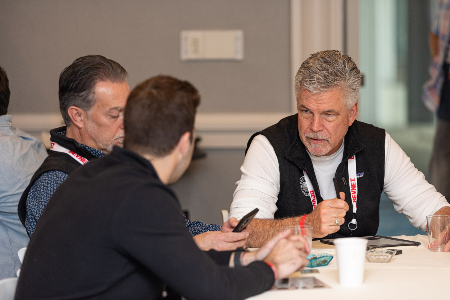 A man in a black vest and BevNET lanyard talks across a table with two other attendees, one holding a phone.