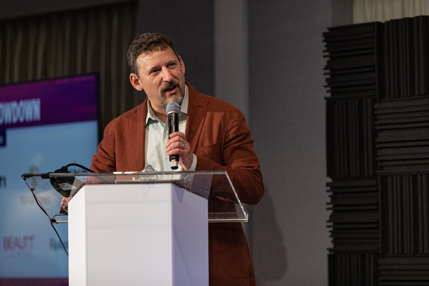 A man in a rust blazer speaks into a handheld microphone at a clear podium on a stage with acoustic foam panels.
