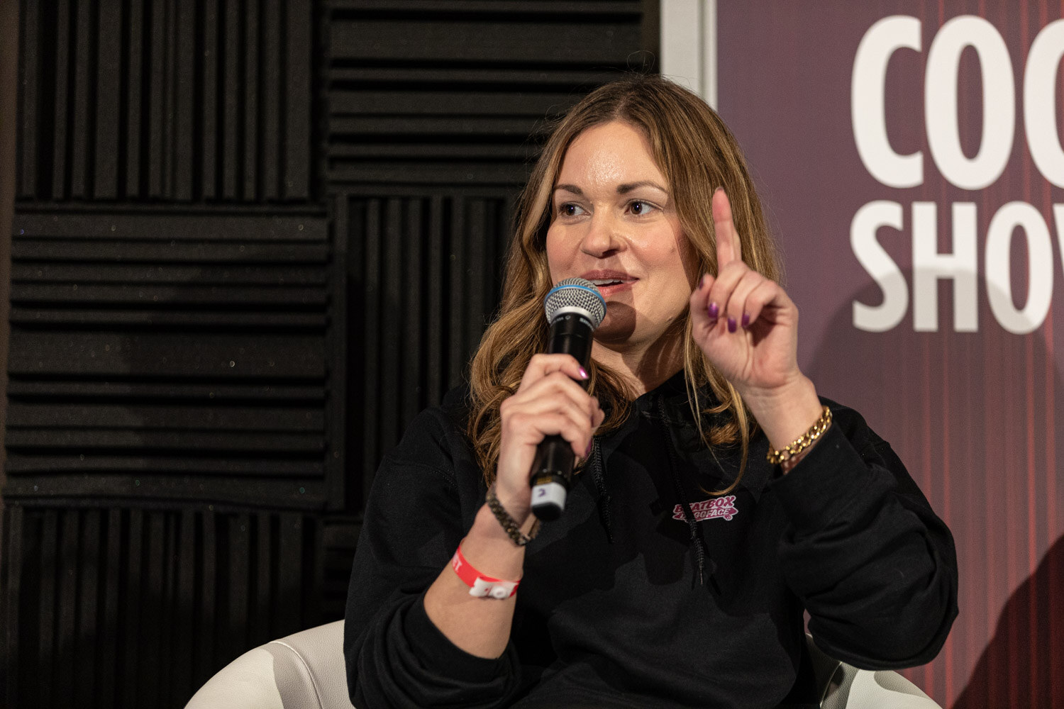 A woman in a black hooded sweatshirt holds a microphone and raises one finger in front of a Cocktail Showdown banner.