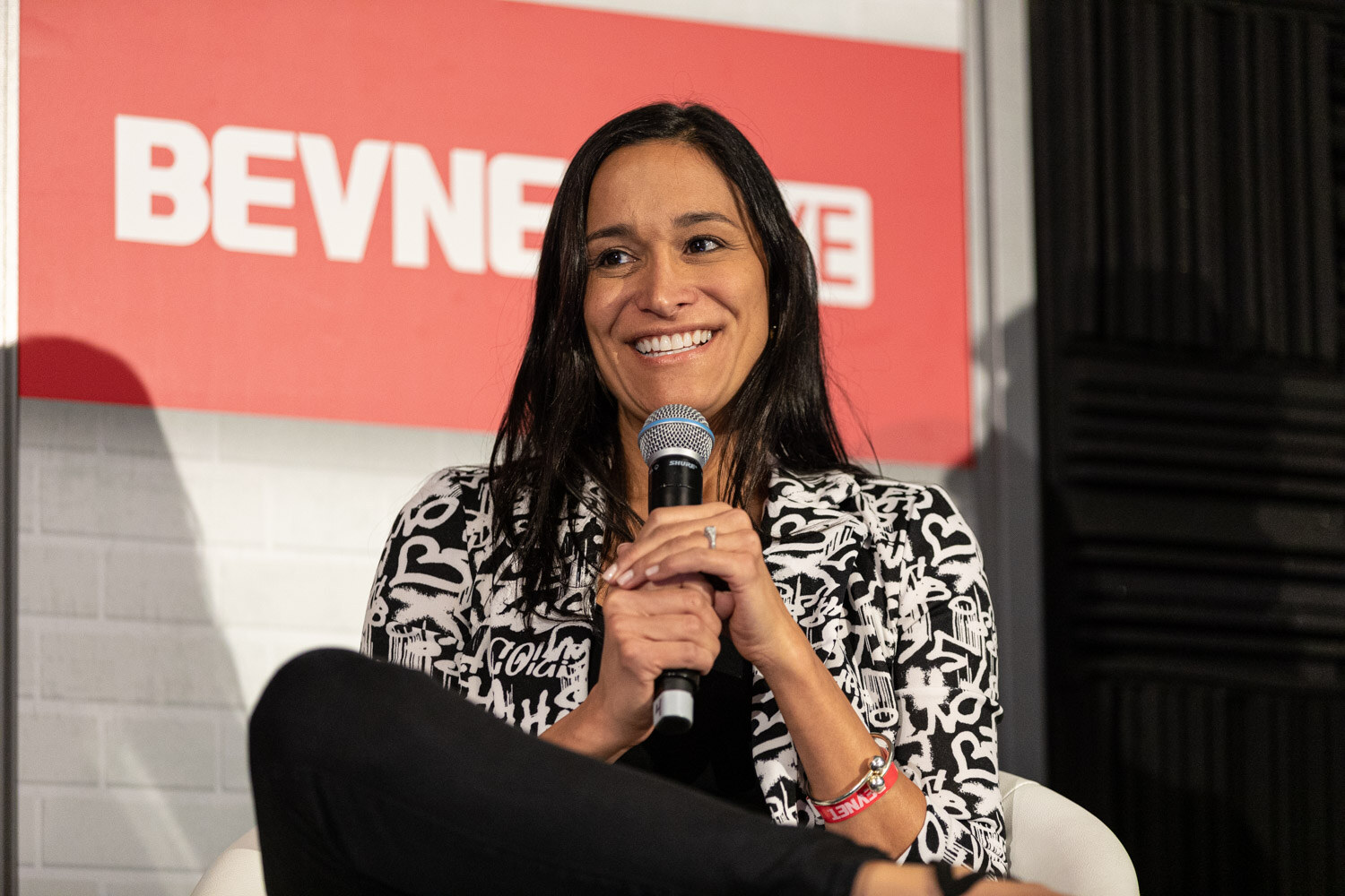 A smiling woman with long dark hair in a black and white patterned jacket holds a microphone in front of a BevNET Live sign.