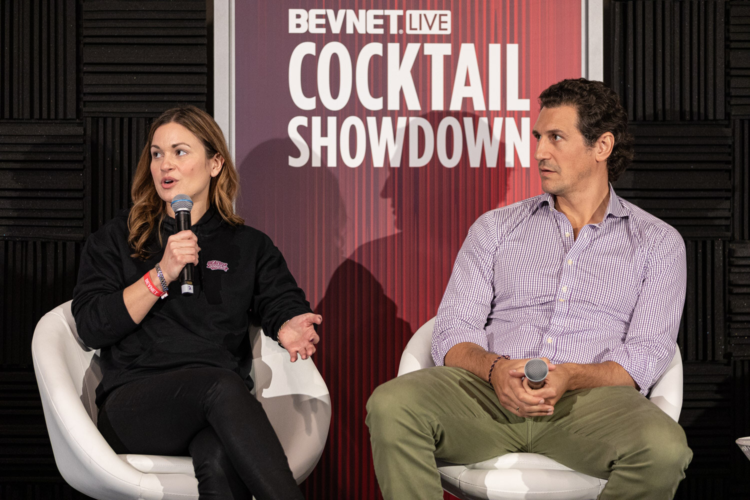 A woman in a black hooded sweatshirt and a man in a lavender shirt and green pants sit in front of a banner reading BevNET Live Cocktail Showdown.