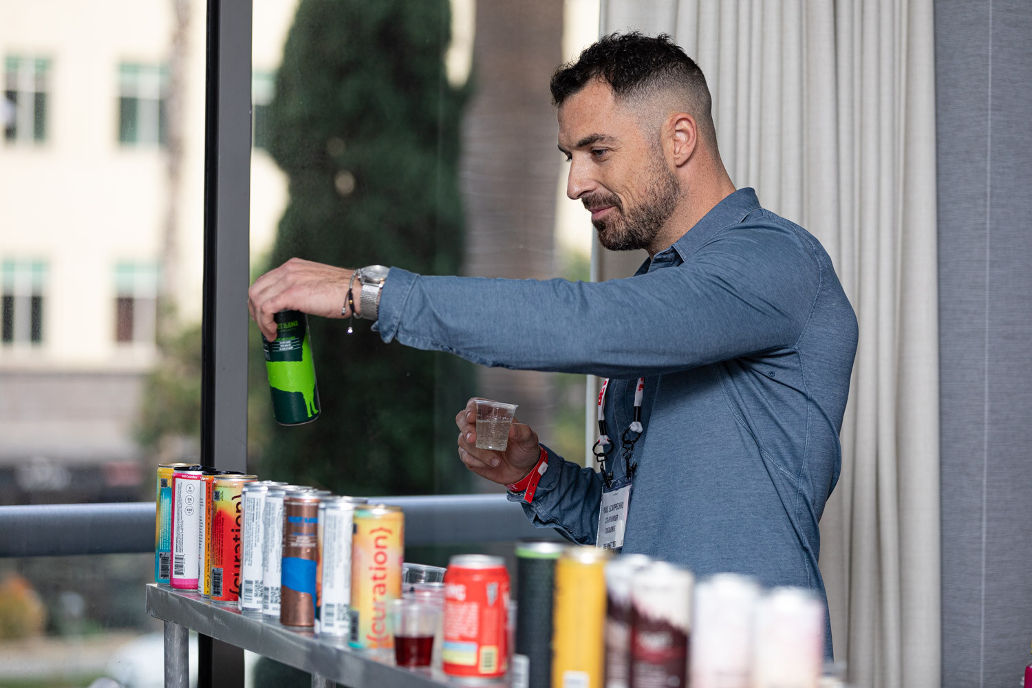 A man in a blue button-up shirt pours a green can into a small cup at a counter lined with canned cocktails in front of a window.