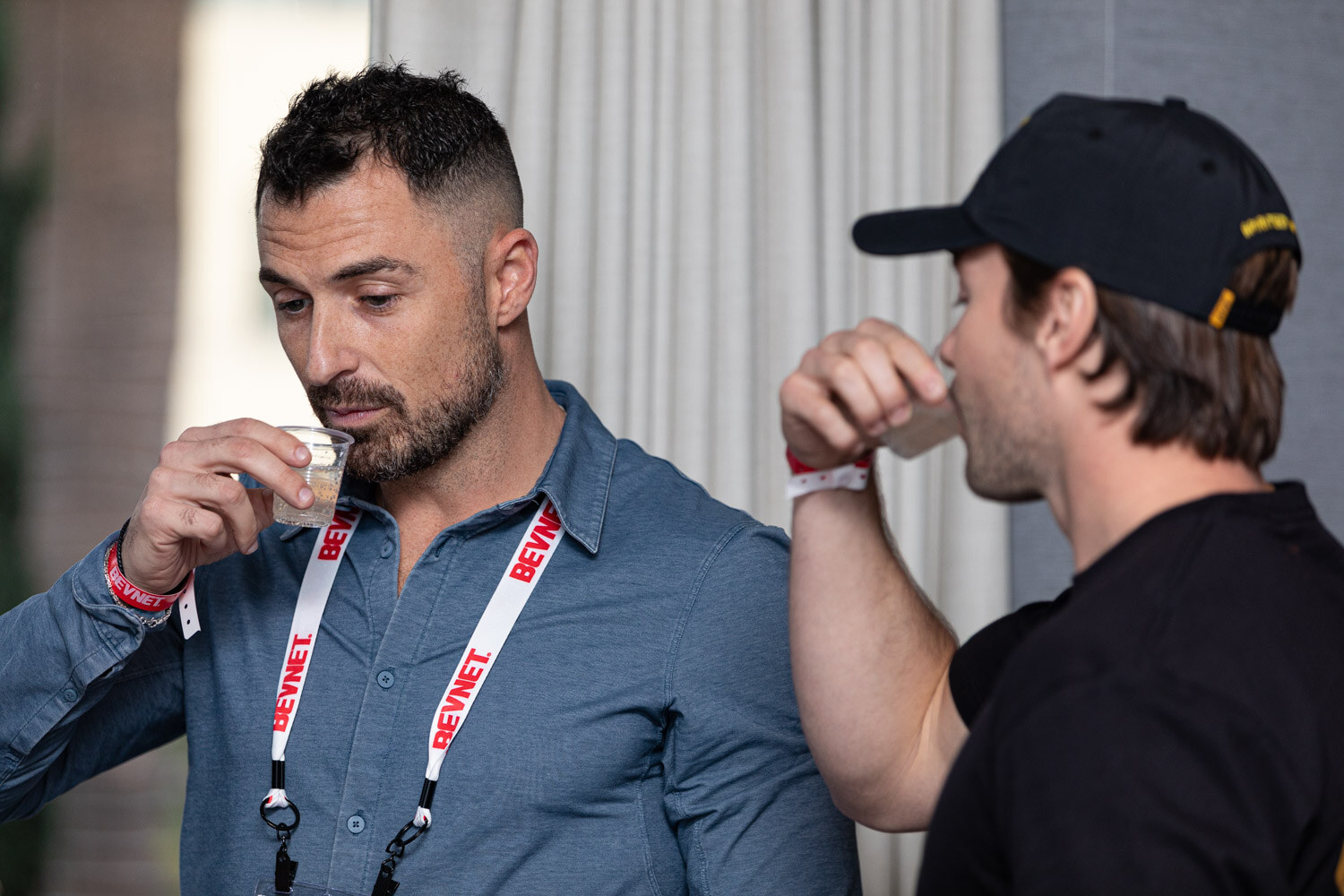 A man with a BevNET lanyard sips from a small cup while a man in a black cap beside him also drinks.