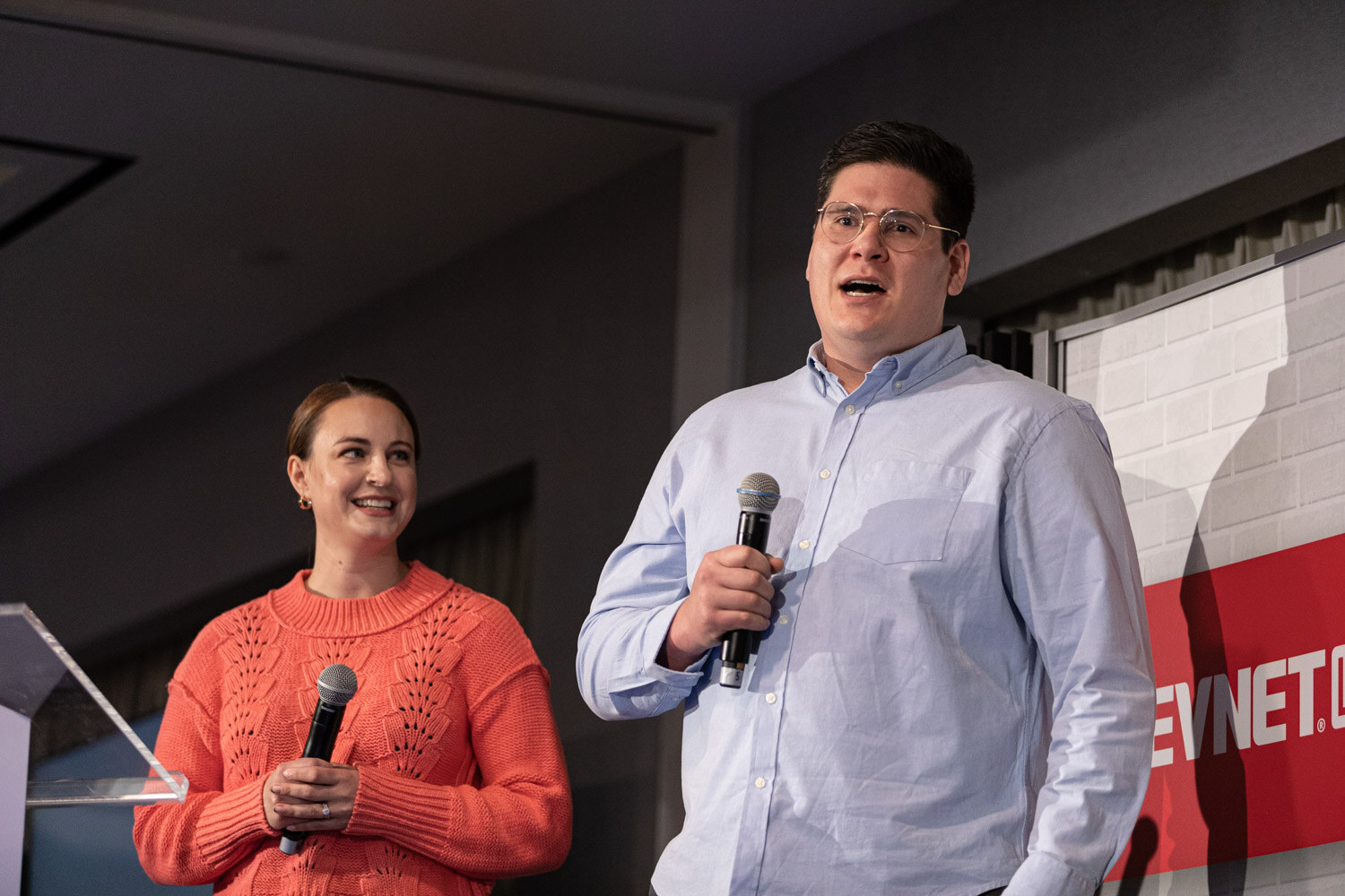 A woman in an orange knit sweater and a man in a light blue shirt hold microphones on a stage next to a clear podium and a BevNET Live sign.