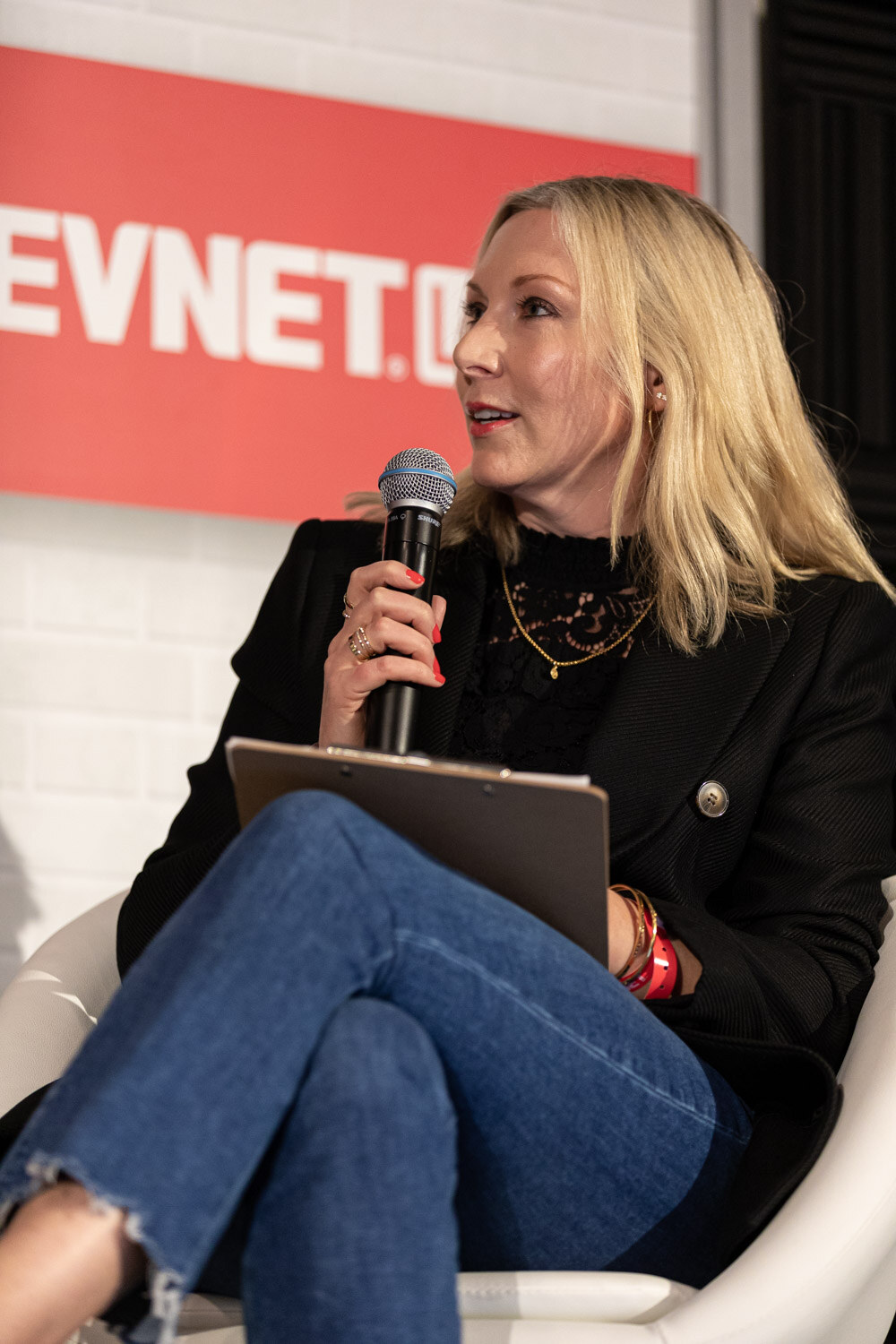 A blond woman in a black blazer and jeans sits with her legs crossed and a clipboard on her knee while holding a microphone.