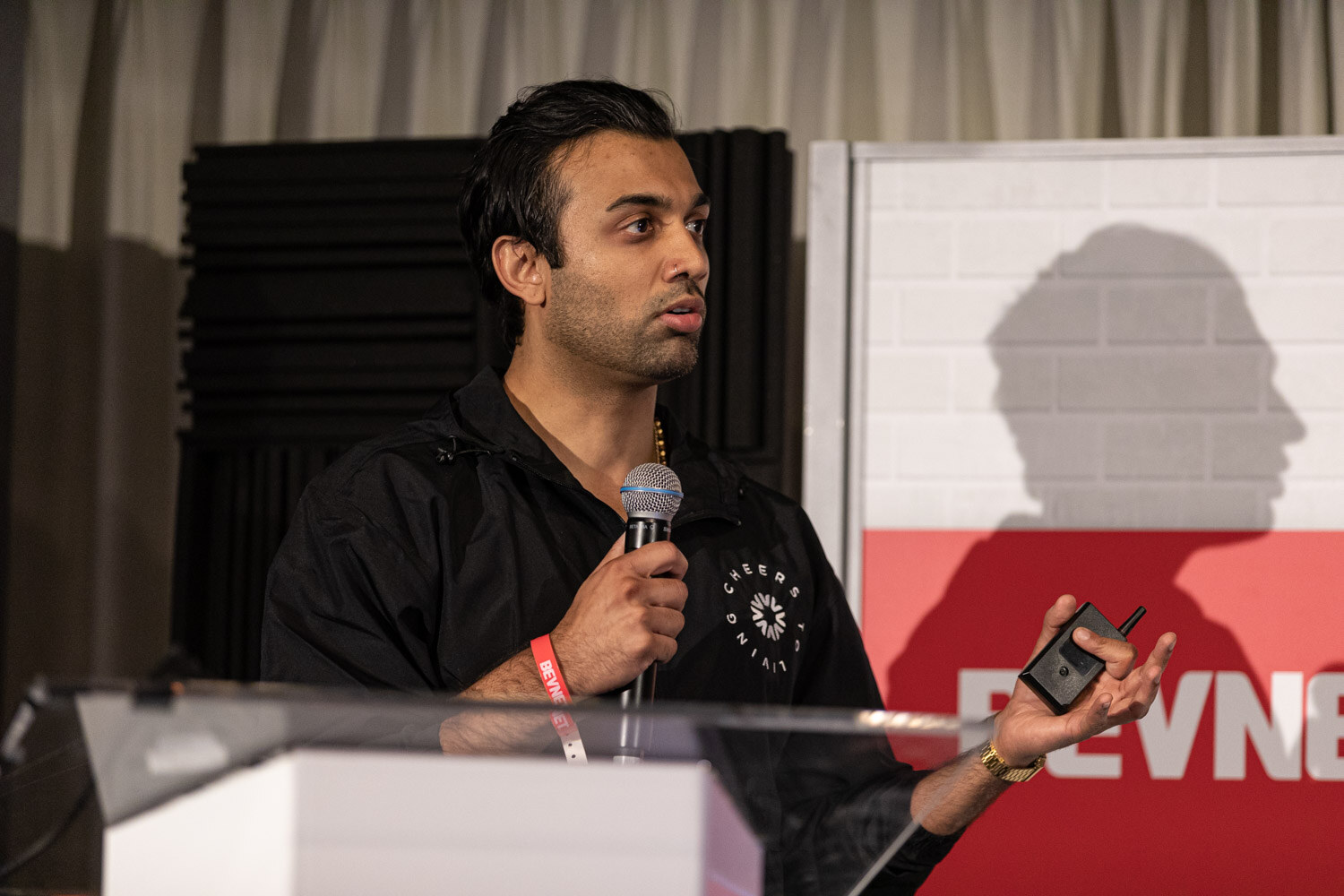 A man in a black zip jacket with a Cheers logo holds a microphone and a clicker behind a clear podium in front of a BevNET sign.