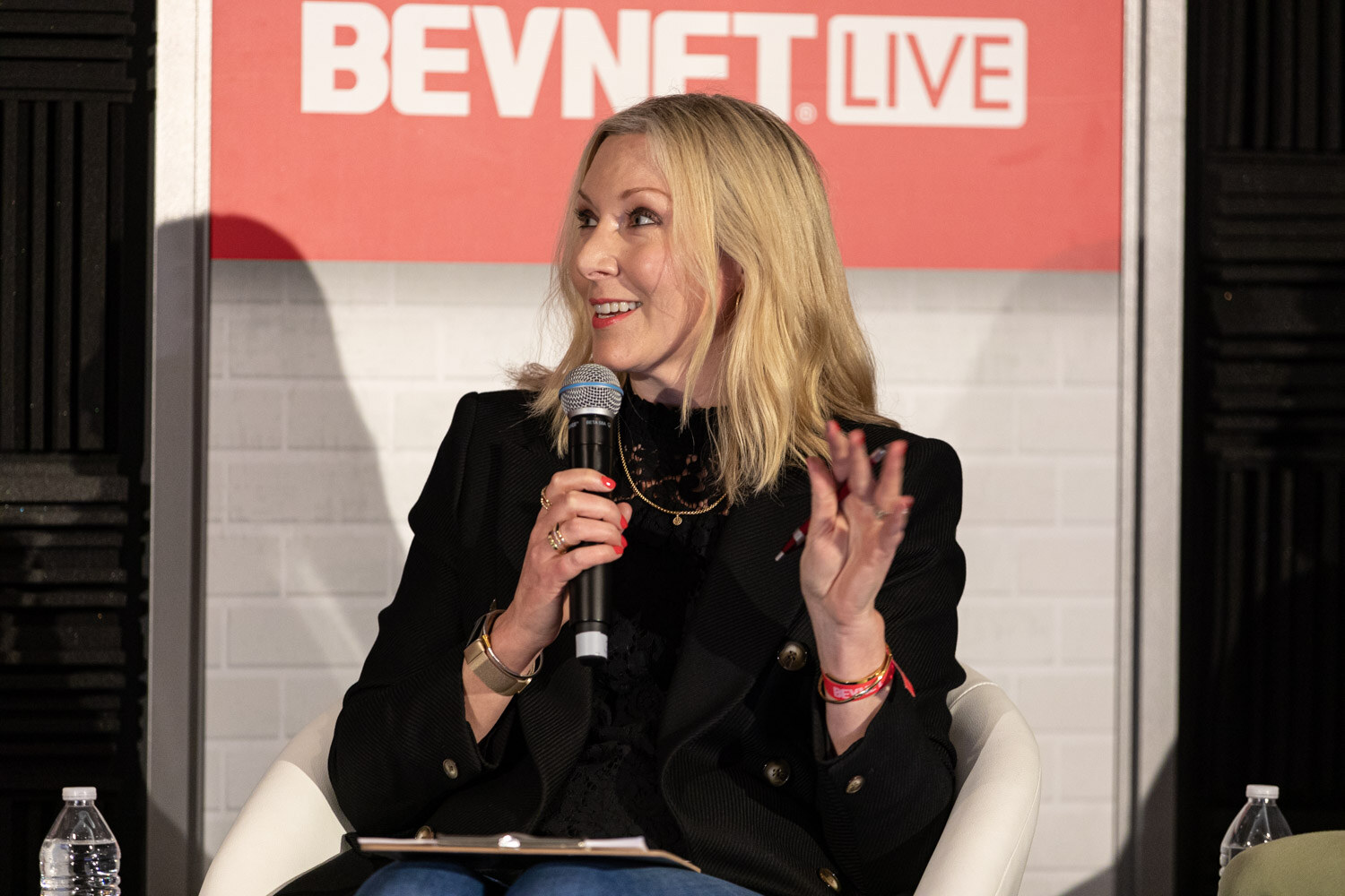 A blond woman in a black blazer holds a microphone and a clipboard as she gestures with one hand in a white chair.
