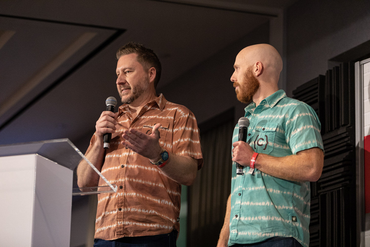 Two men in tie-dye style short-sleeved shirts hold microphones on stage behind a clear podium, one speaking and the other looking at him.