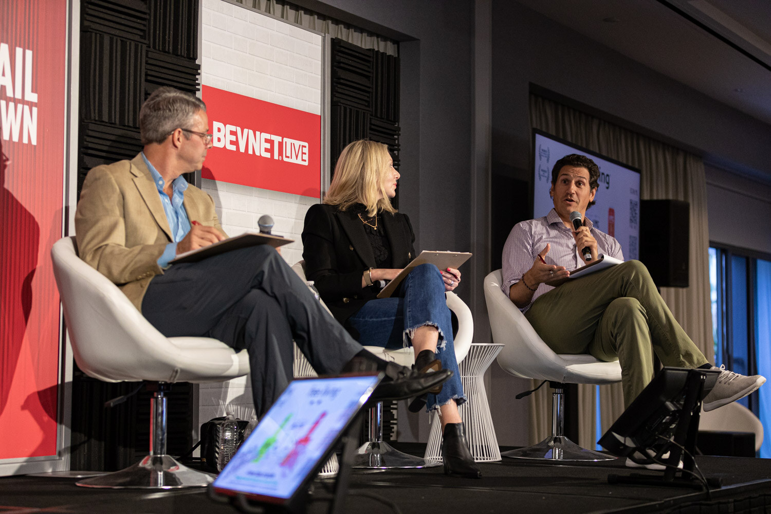 A man in a tan blazer, a blond woman in a black blazer and a man in a lavender shirt sit in white chairs in front of BevNET Live signs, holding clipboards.