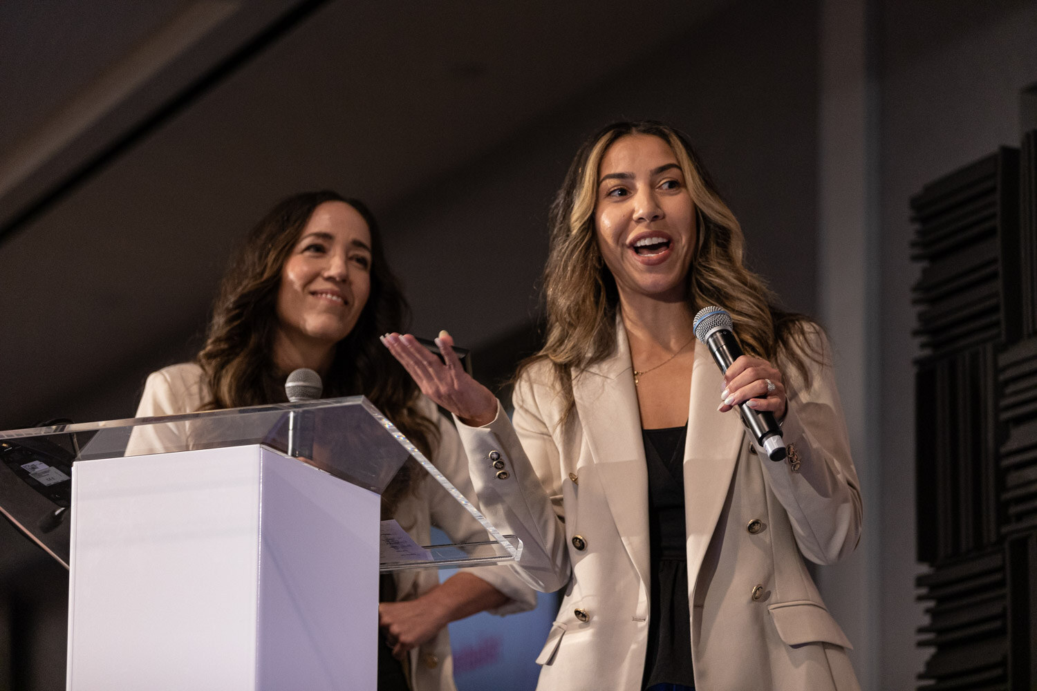 Two women in cream blazers stand behind a clear podium, one holding a microphone and speaking with a bright expression.