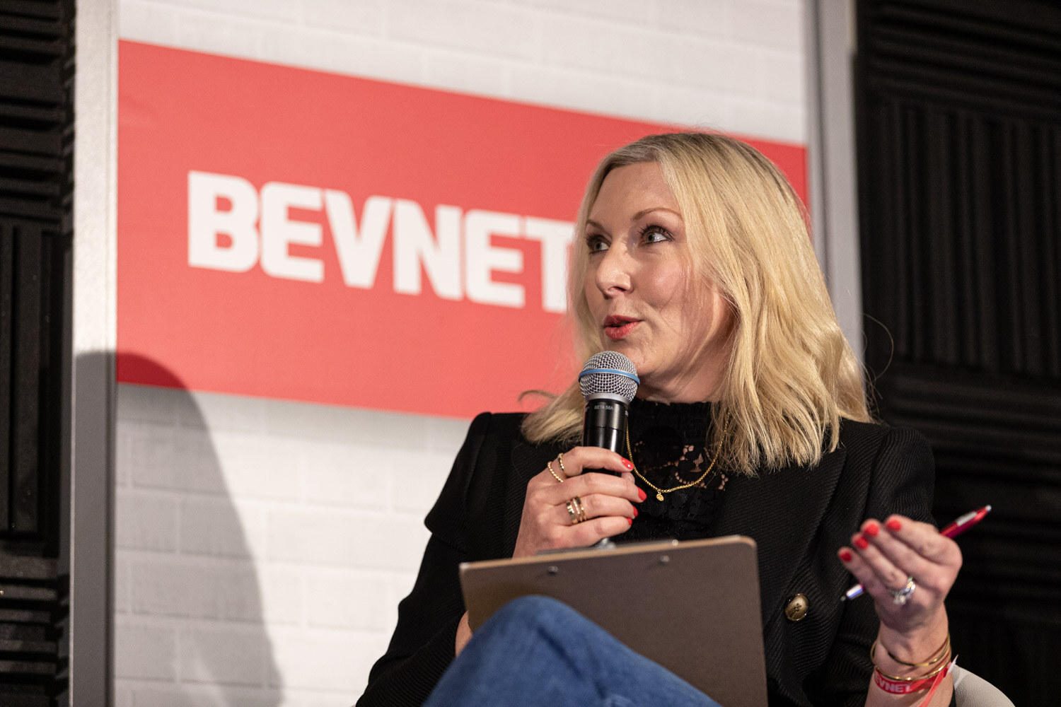 A blond woman in a black blazer sits in front of a red BevNET sign holding a microphone, a clipboard and a pen.