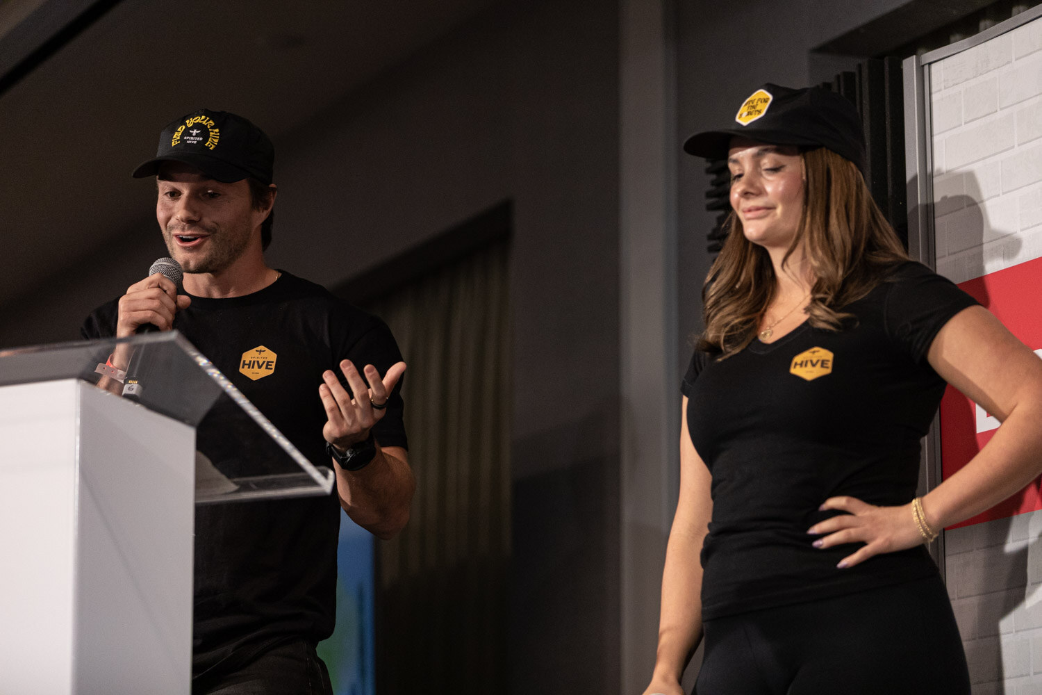 A man and a woman in black t-shirts and caps with a yellow hexagon Hive logo stand behind a clear podium, with the man holding a microphone and gesturing.