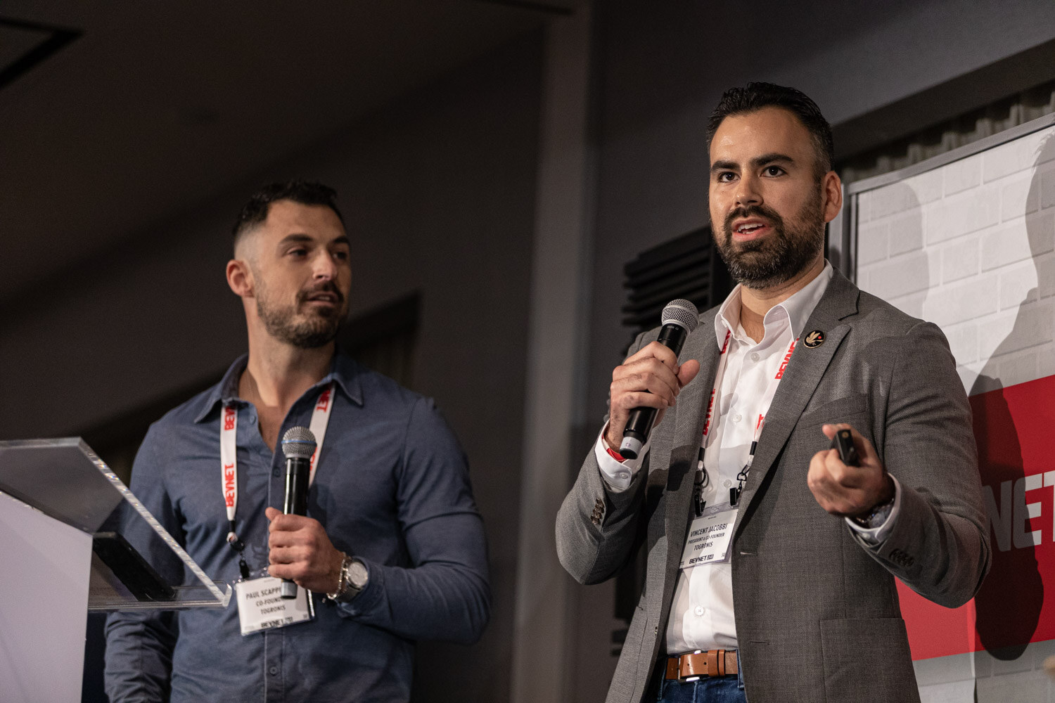 A man in a blue button-up and a man in a gray blazer both hold microphones at a clear podium, with the second man holding a clicker.
