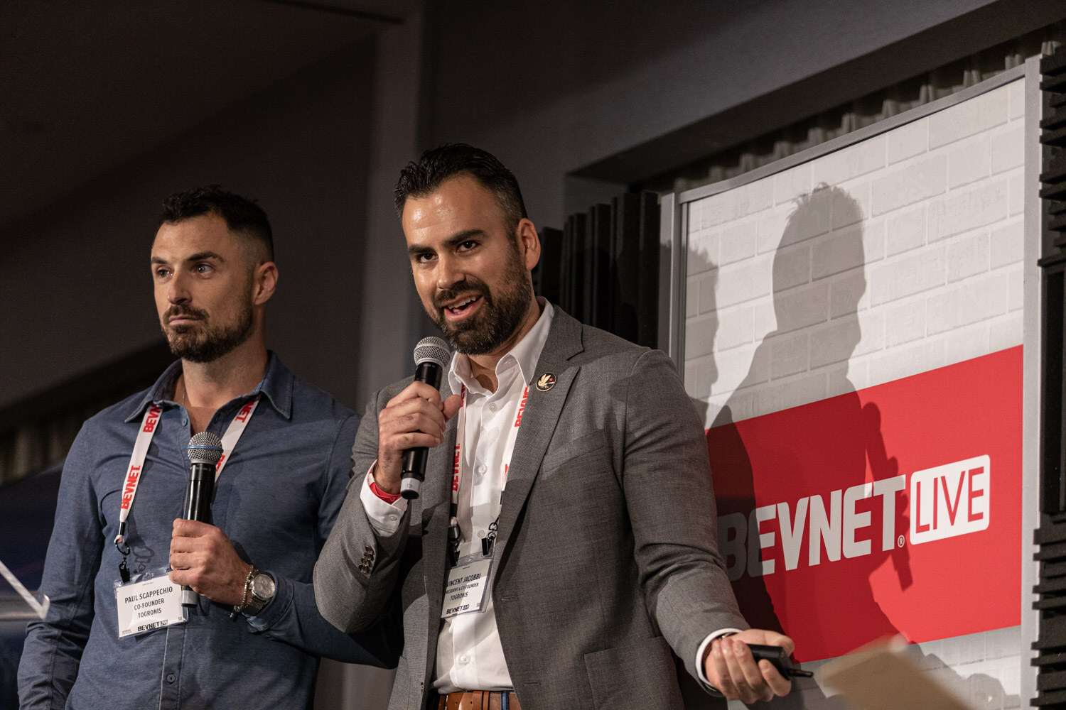 Two men with BevNET lanyards stand in front of a BevNET Live sign, one holding a microphone and speaking, the other looking on.