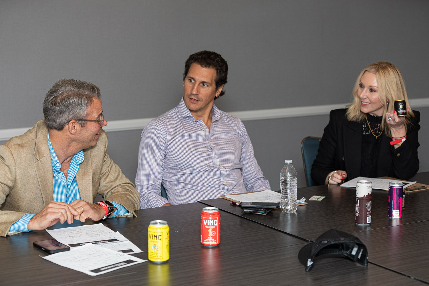 Two men and a blond woman sit at a long table with scoring sheets and canned cocktails including cans reading Ving.