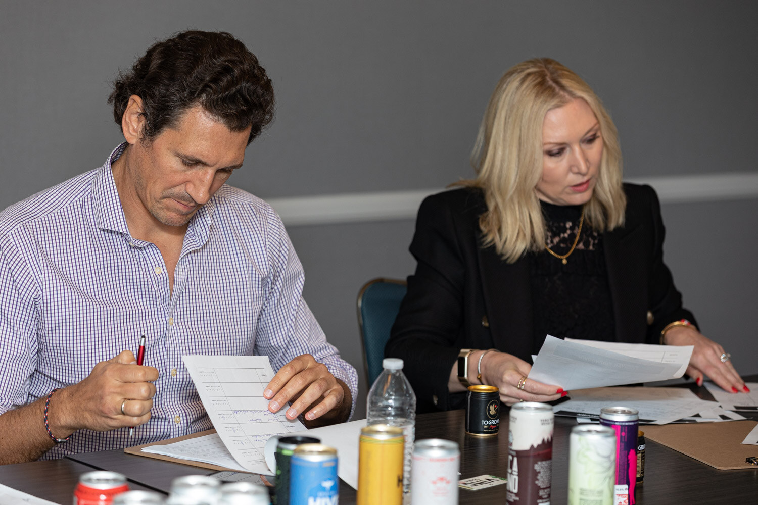 A man in a lavender shirt holding a pen and a blond woman in a black blazer review scoring sheets next to a row of canned cocktails.