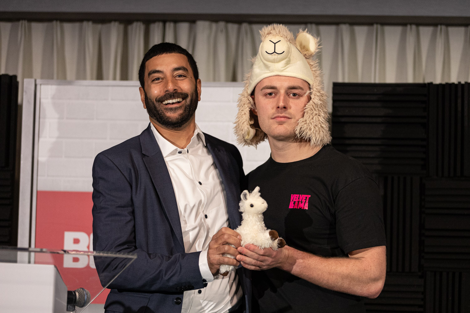 A smiling man in a navy blazer and a man in a black t-shirt wearing a plush llama hat hold a white llama toy together on stage.