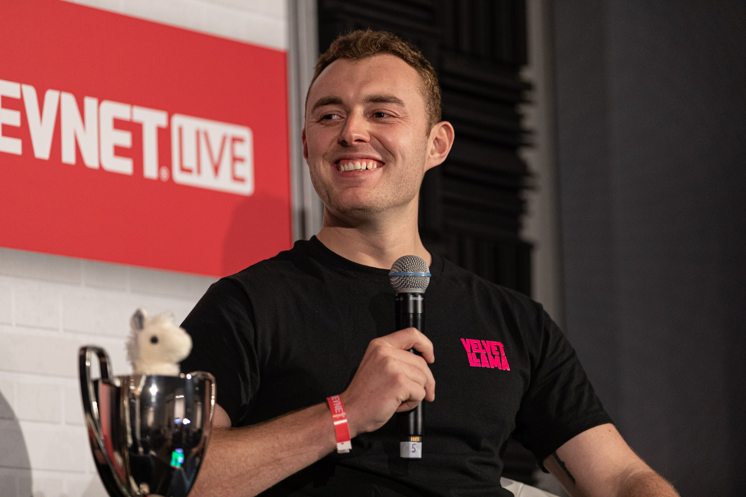 A smiling young man in a black Velvet Llama t-shirt holds a microphone and looks off to his left, with a trophy and a white llama toy beside him.