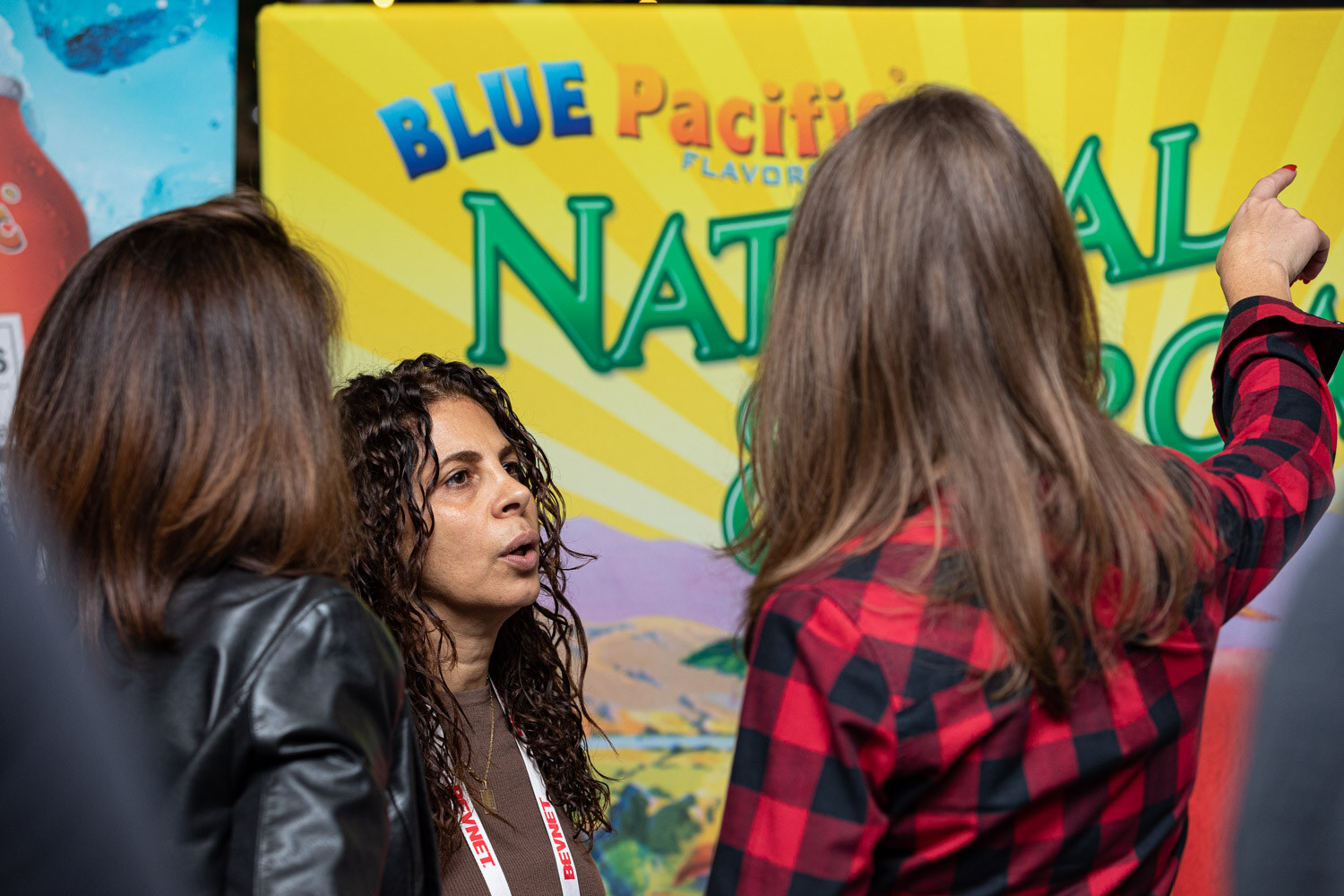A woman with curly hair in a black jacket and a woman in a red plaid shirt gesture toward a yellow Blue Pacific Flavors banner.