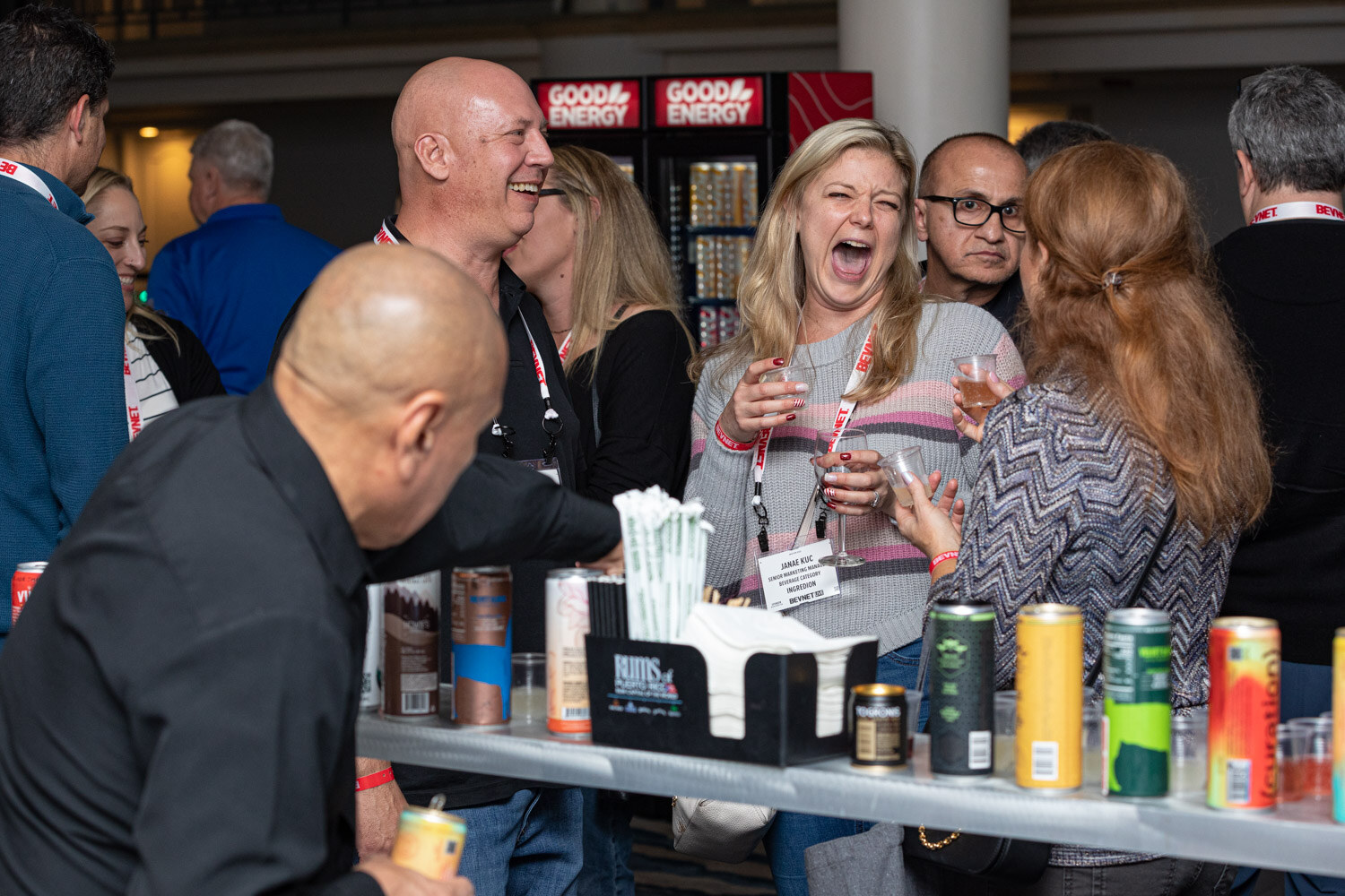 A group of attendees stand around a table lined with cans and cups while a woman in a striped sweater laughs with her mouth open.