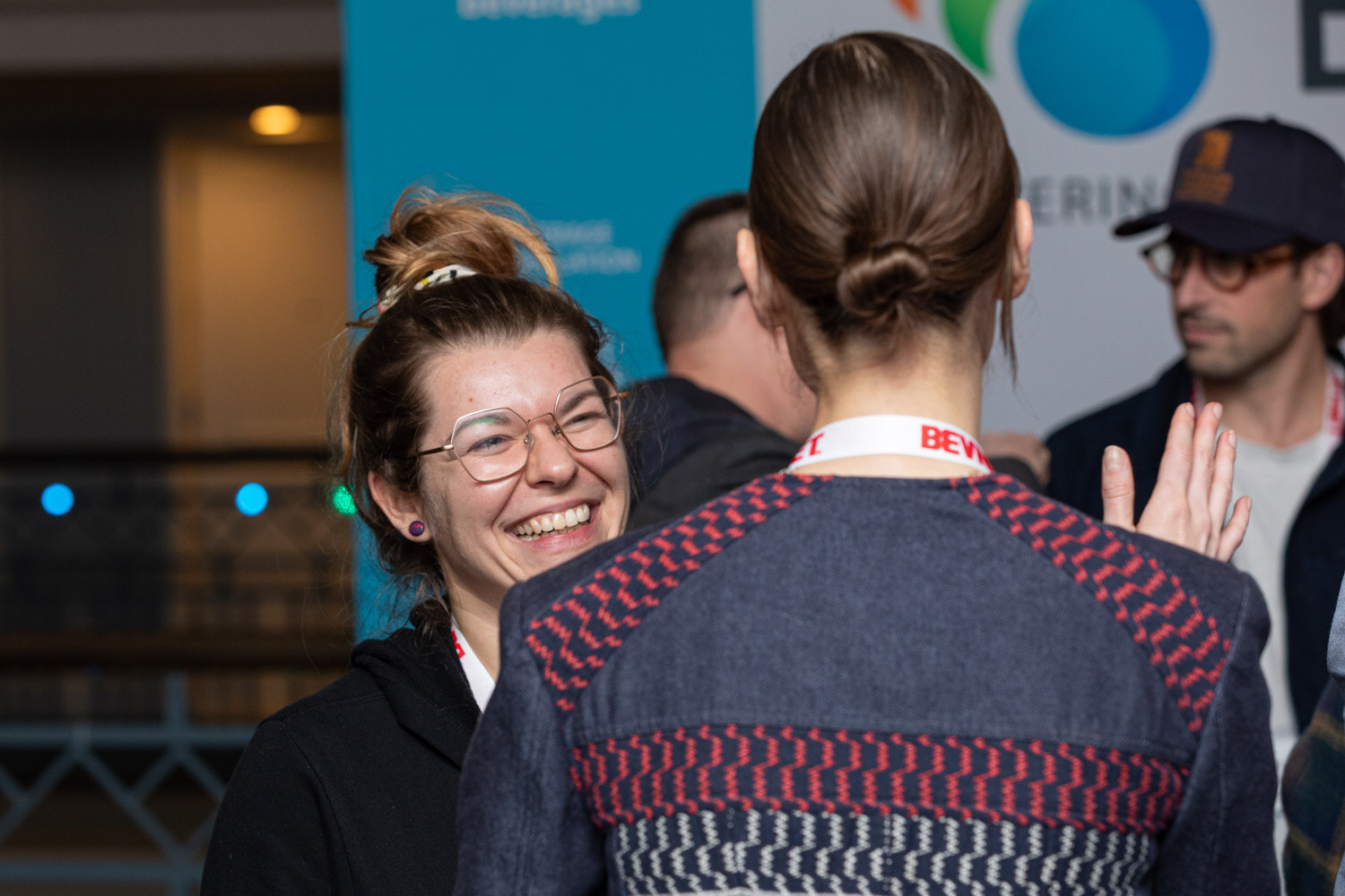 A woman with glasses and a scrunchie-tied bun laughs as she talks with a woman seen from behind in a patterned sweater with a BevNET lanyard.