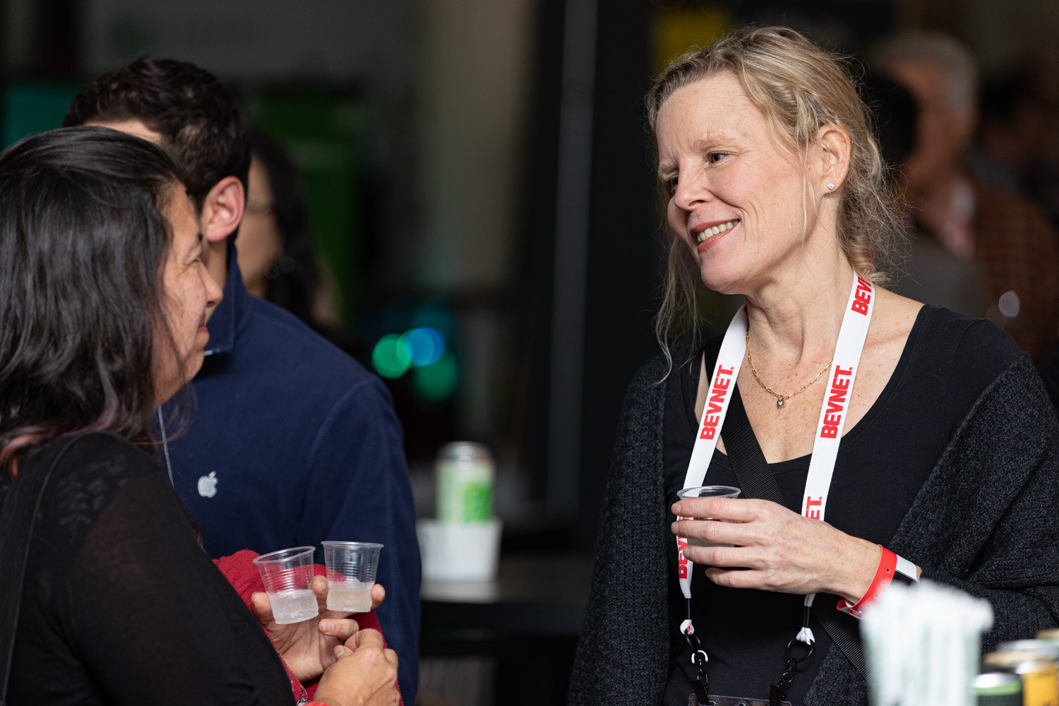 A blond woman in black with a BevNET lanyard and a plastic sample cup talks with a woman holding two cups in front of a dark background.