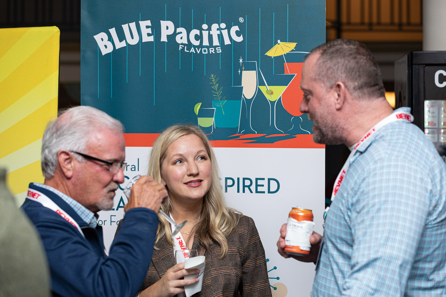 A woman in a tweed jacket stands between two men, one in a plaid shirt holding a canned drink, in front of a Blue Pacific Flavors banner.