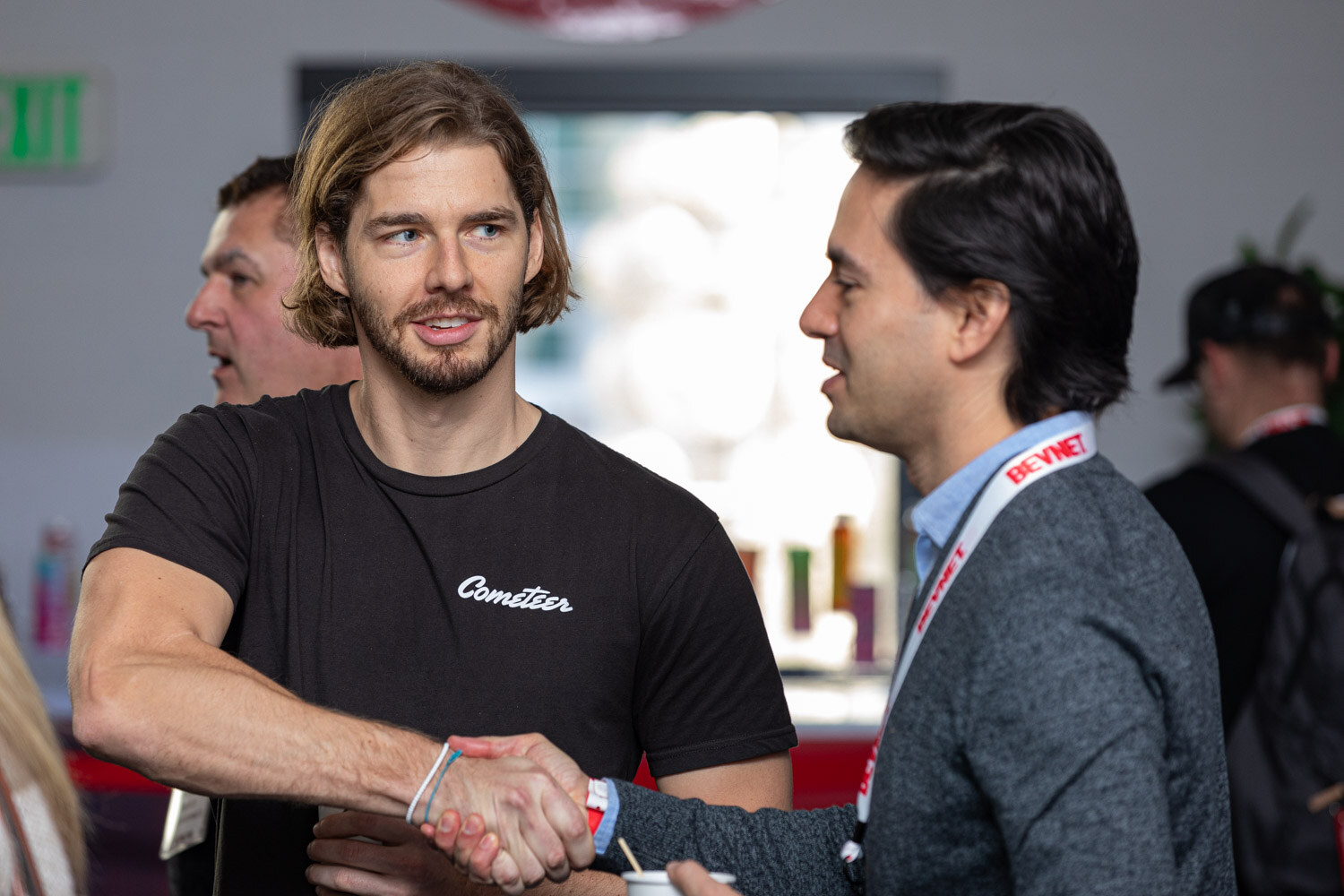A man with shoulder-length hair in a black Cometeer t-shirt shakes hands with a man in a gray sweater wearing a BevNET lanyard.