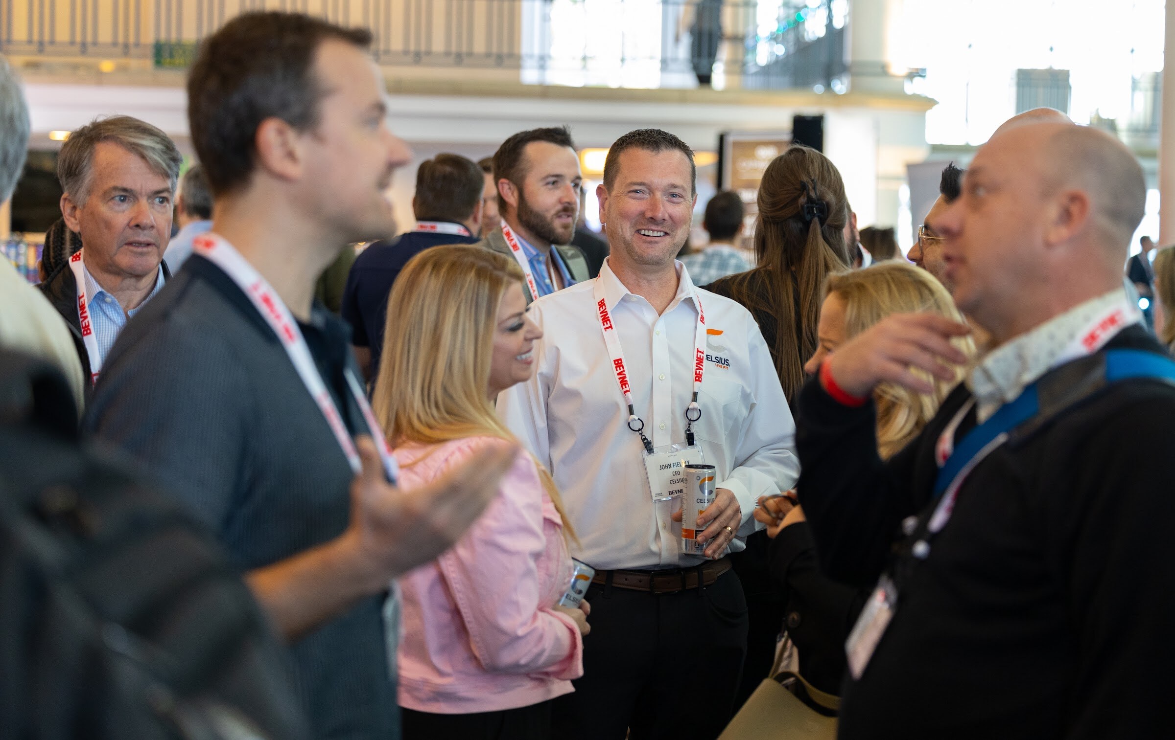 A group of attendees wearing BevNET lanyards chat in a bright hotel atrium, with a man in a white shirt smiling in the center.