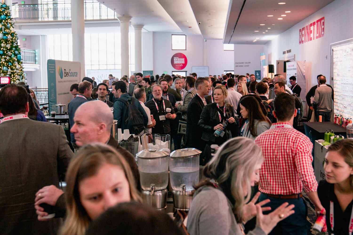 A crowd of attendees stands around beverage stations in a bright atrium with a decorated Christmas tree and a BevNET Live sign.
