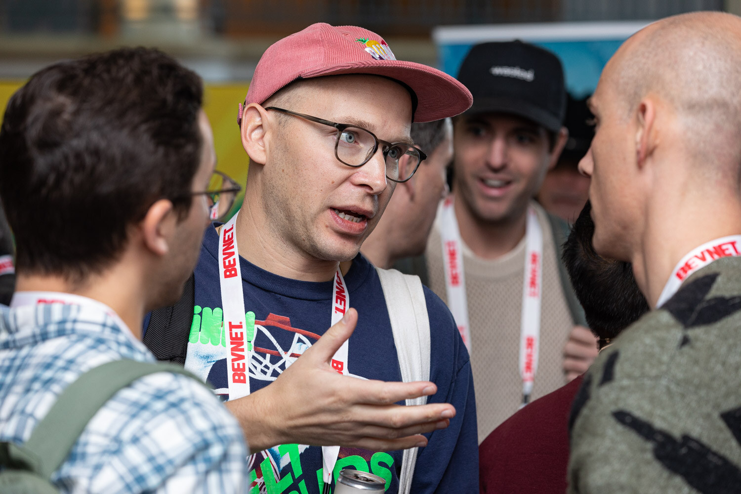 A man in a pink baseball cap and glasses with a BevNET lanyard speaks and gestures to a group of attendees.