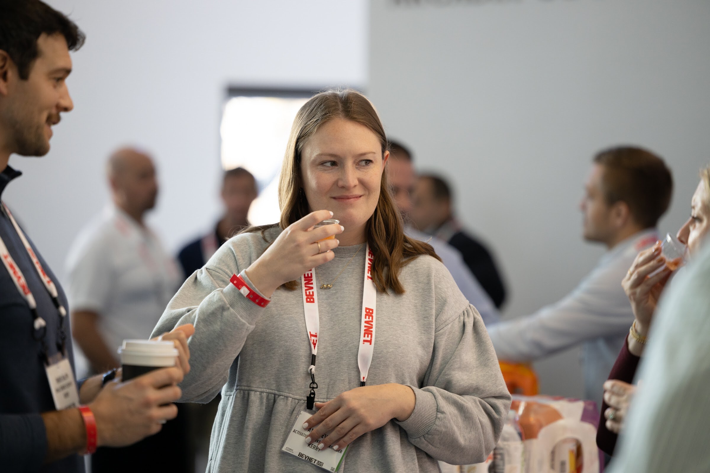A woman in a gray sweatshirt with a BevNET lanyard holds a sample cup to her mouth while others talk behind her.