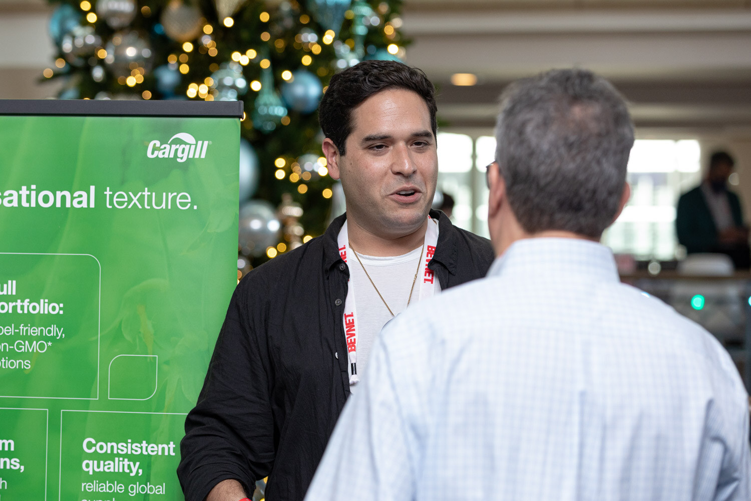 A man in a black shirt with a BevNET lanyard talks with a man in a light checked shirt in front of a Cargill banner and a Christmas tree.
