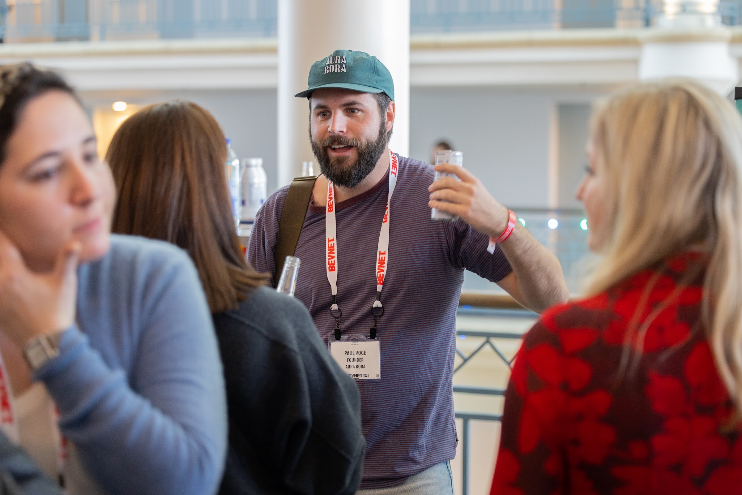 A bearded man in a green cap and a maroon t-shirt with a BevNET lanyard gestures while holding a clear bottle, with other attendees around him.