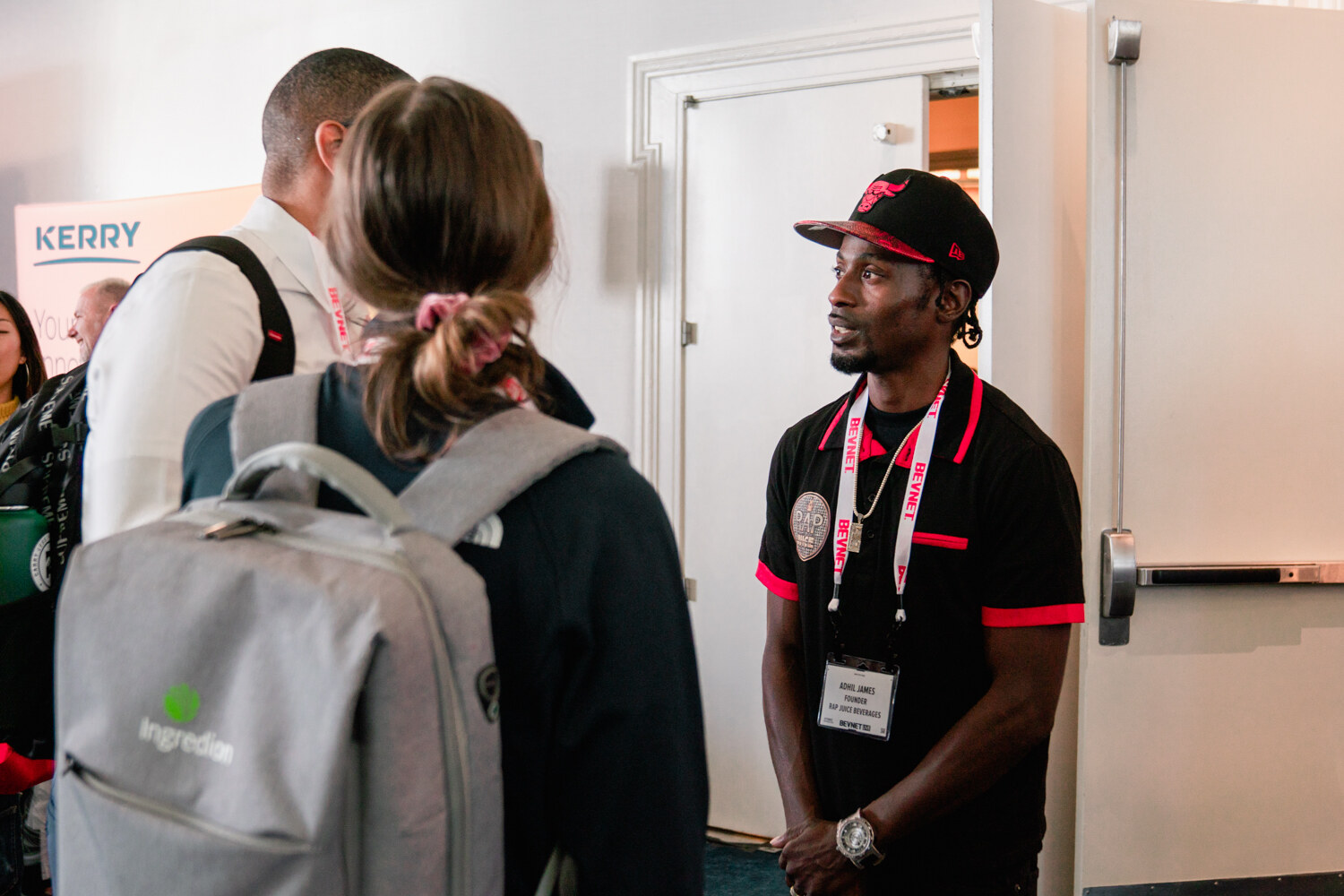 A man in a black and red polo and a red cap with a BevNET lanyard talks with an attendee in a gray Ingredion backpack in a hallway.