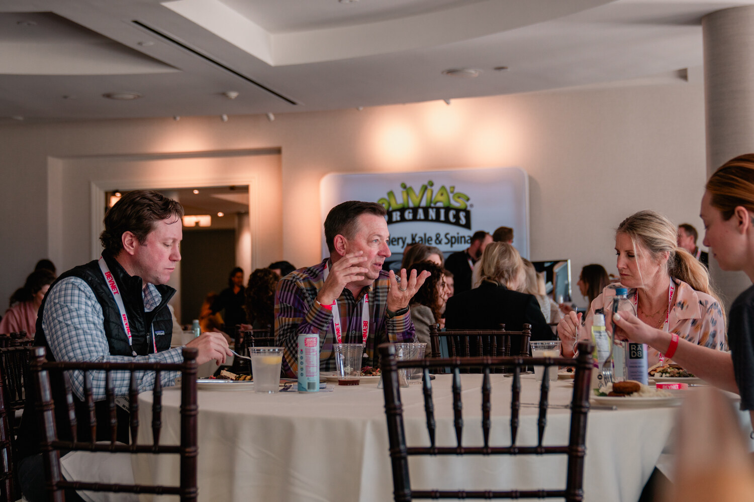 A hotel ballroom with attendees seated at round tables, with an Olivia's Organics banner in the background.