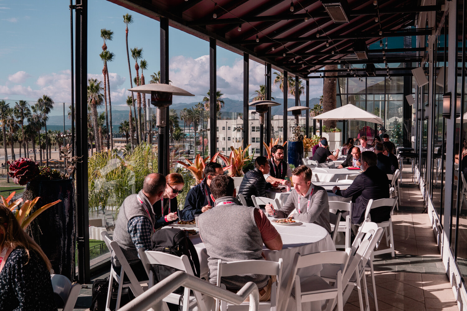 Several tables of attendees eating on an open-sided terrace with palm trees, patio heaters and a hazy coastline in the background.
