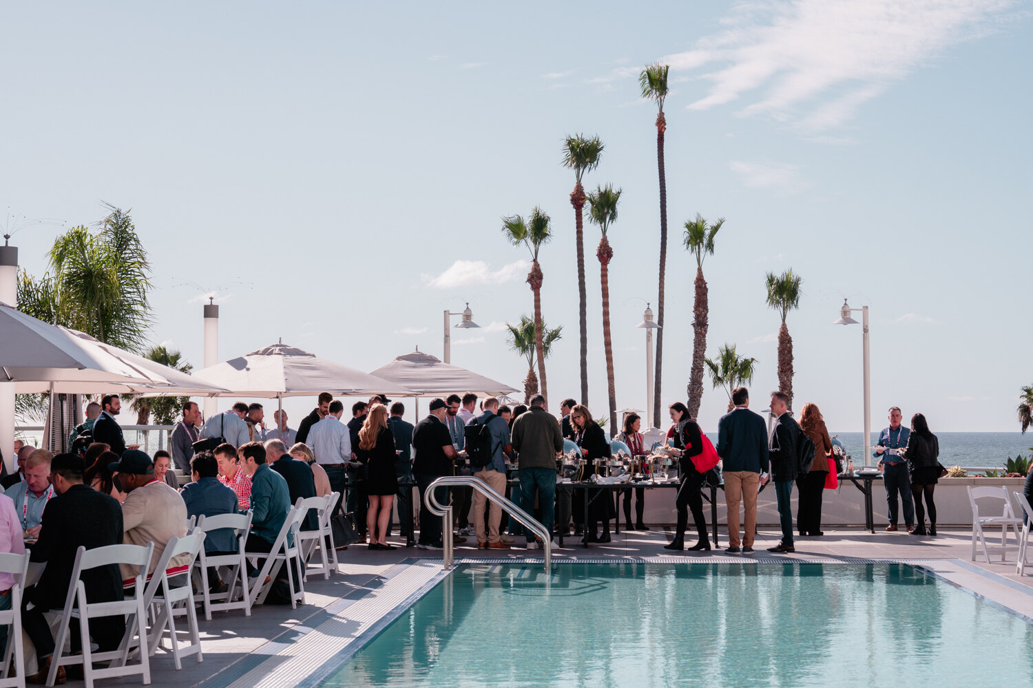 A crowd of attendees at a buffet beside an outdoor pool framed by palm trees and umbrellas under a blue sky.