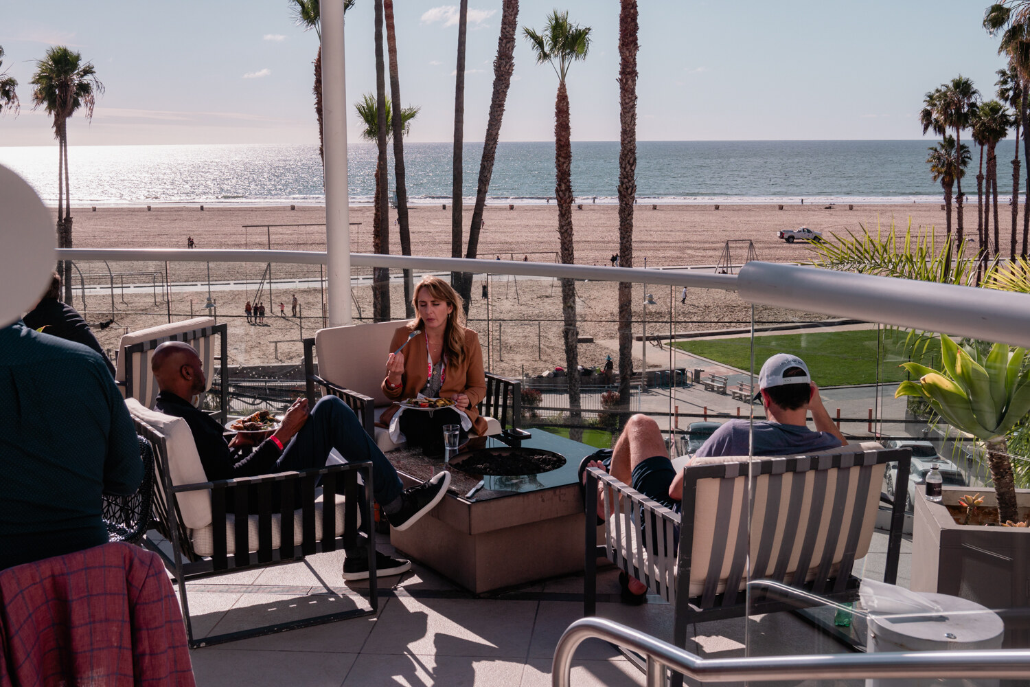 Several people sit and eat on terrace lounge seating next to a glass railing, with palm trees, sand and the ocean in the background.