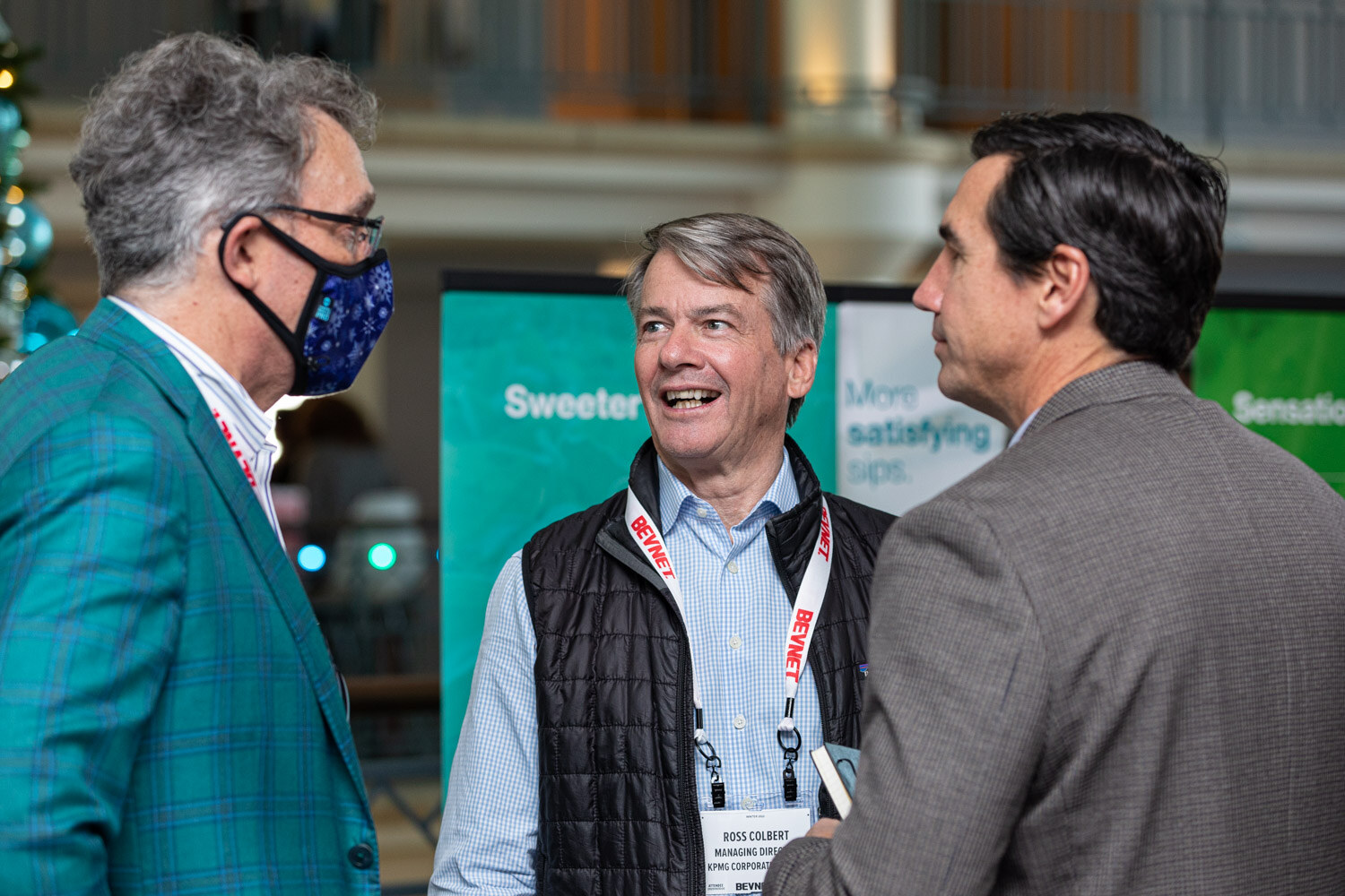 A man in a teal plaid blazer wearing a face mask, a smiling man in a black vest and a man in a gray blazer talk in front of a green display screen.