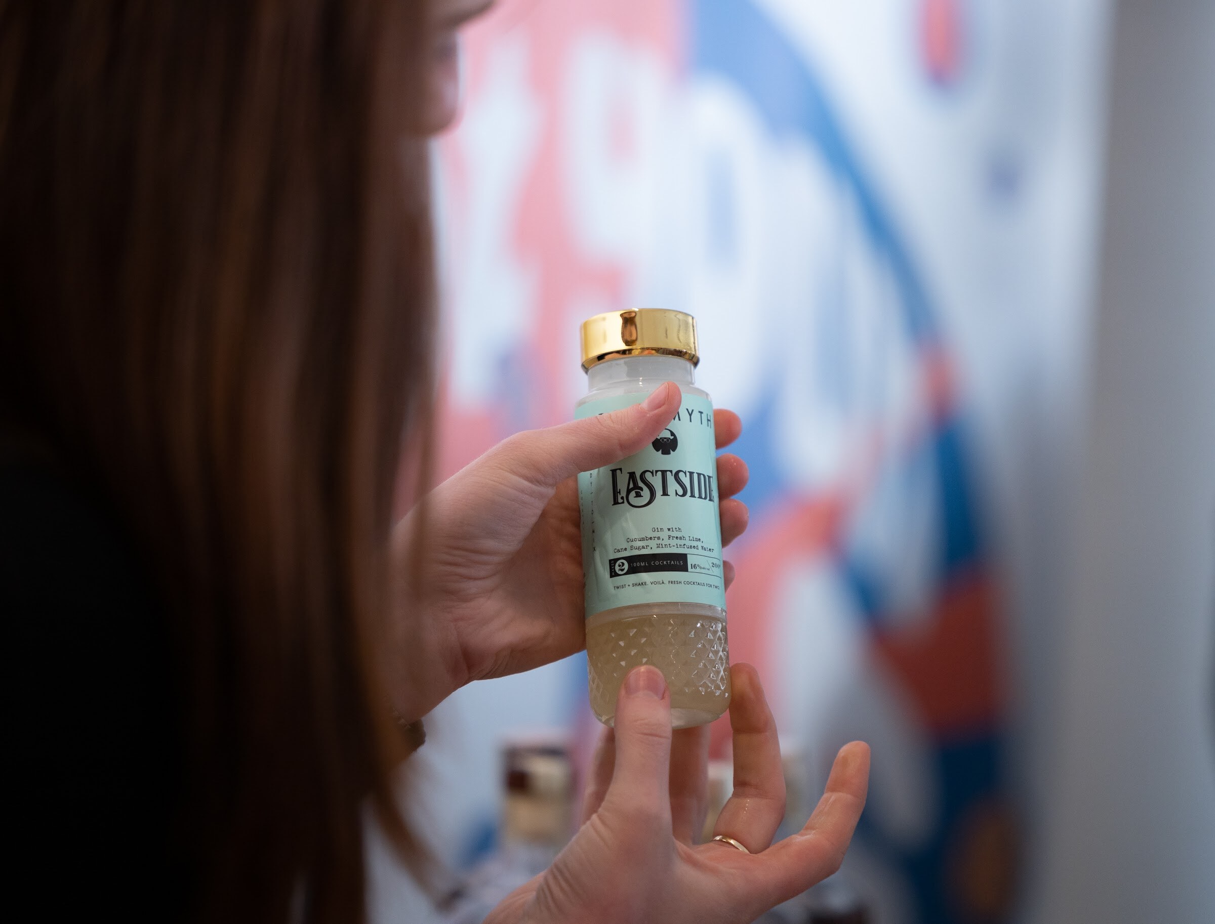 A woman's hands hold a small clear bottle with a gold cap and the label Eastside in front of a blurred red, white and blue banner.