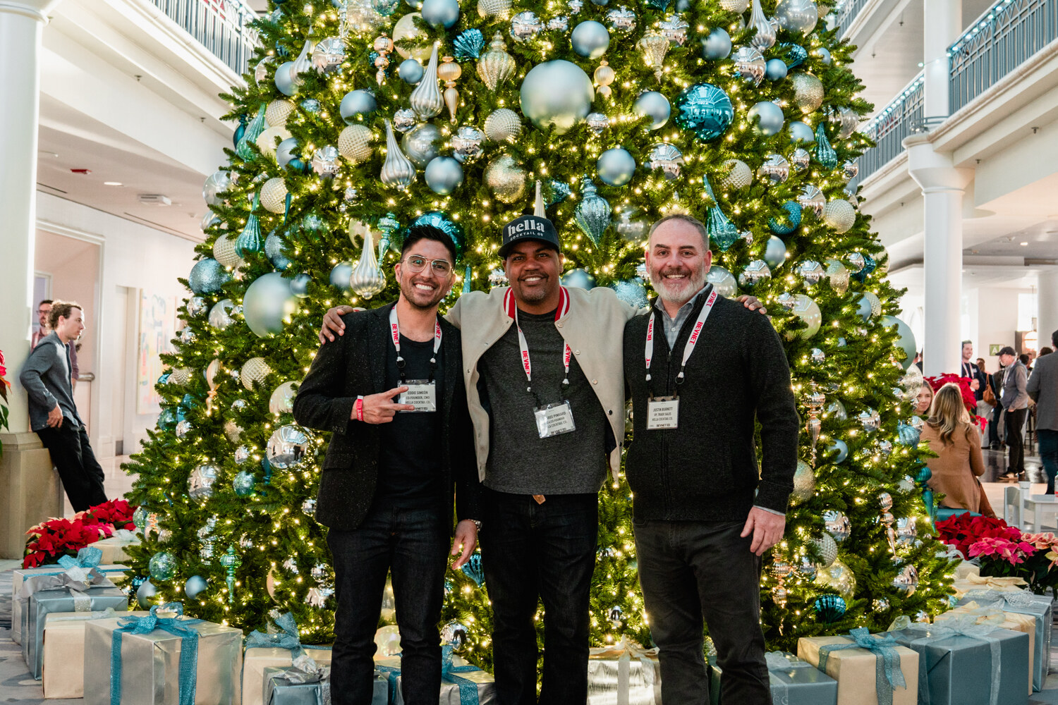 Three men with BevNET lanyards stand arm in arm in front of a tall Christmas tree decorated with blue and silver ornaments.