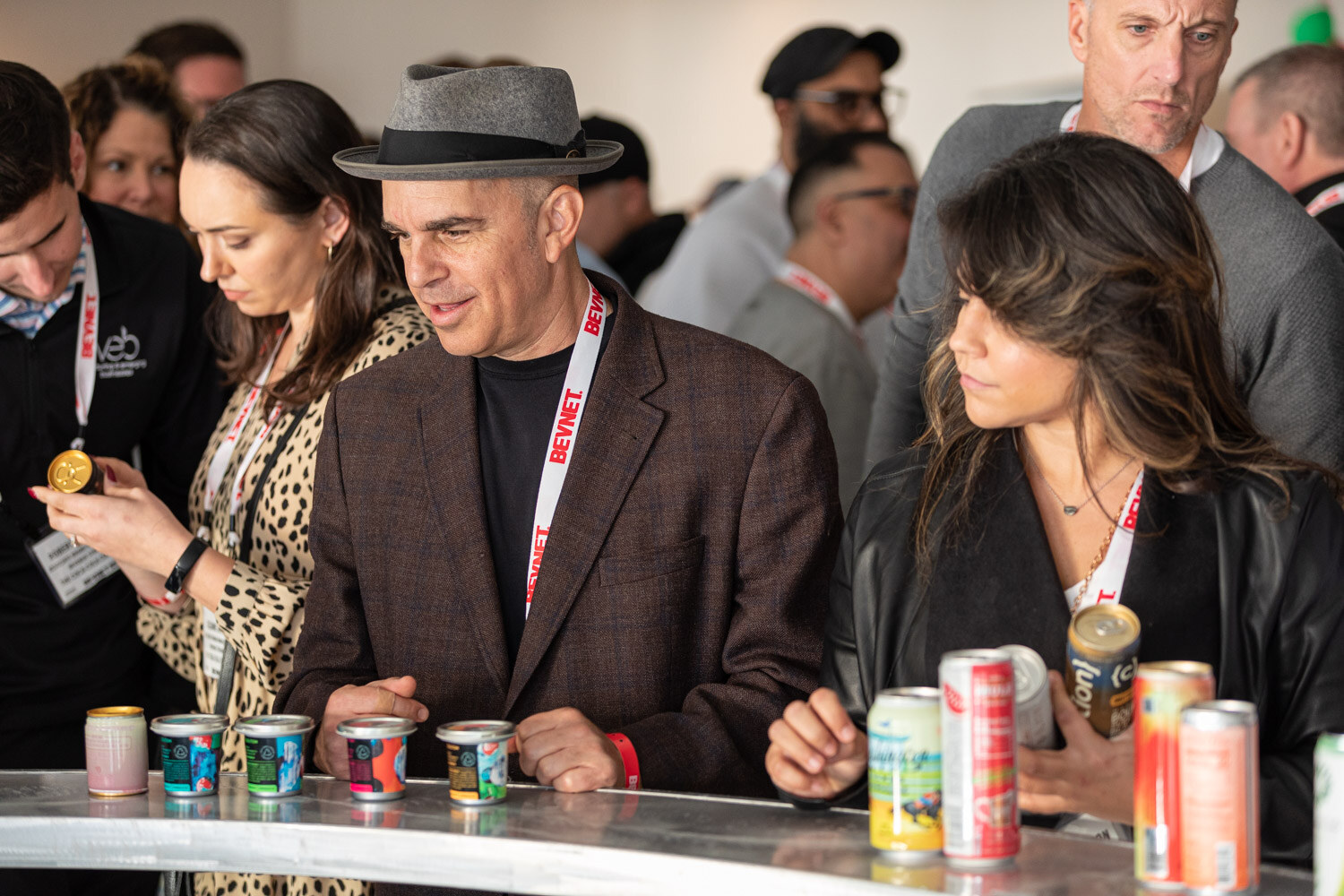 A man in a gray hat and a brown blazer and a woman with long dark hair pick up cans from a counter covered with colorful cans.