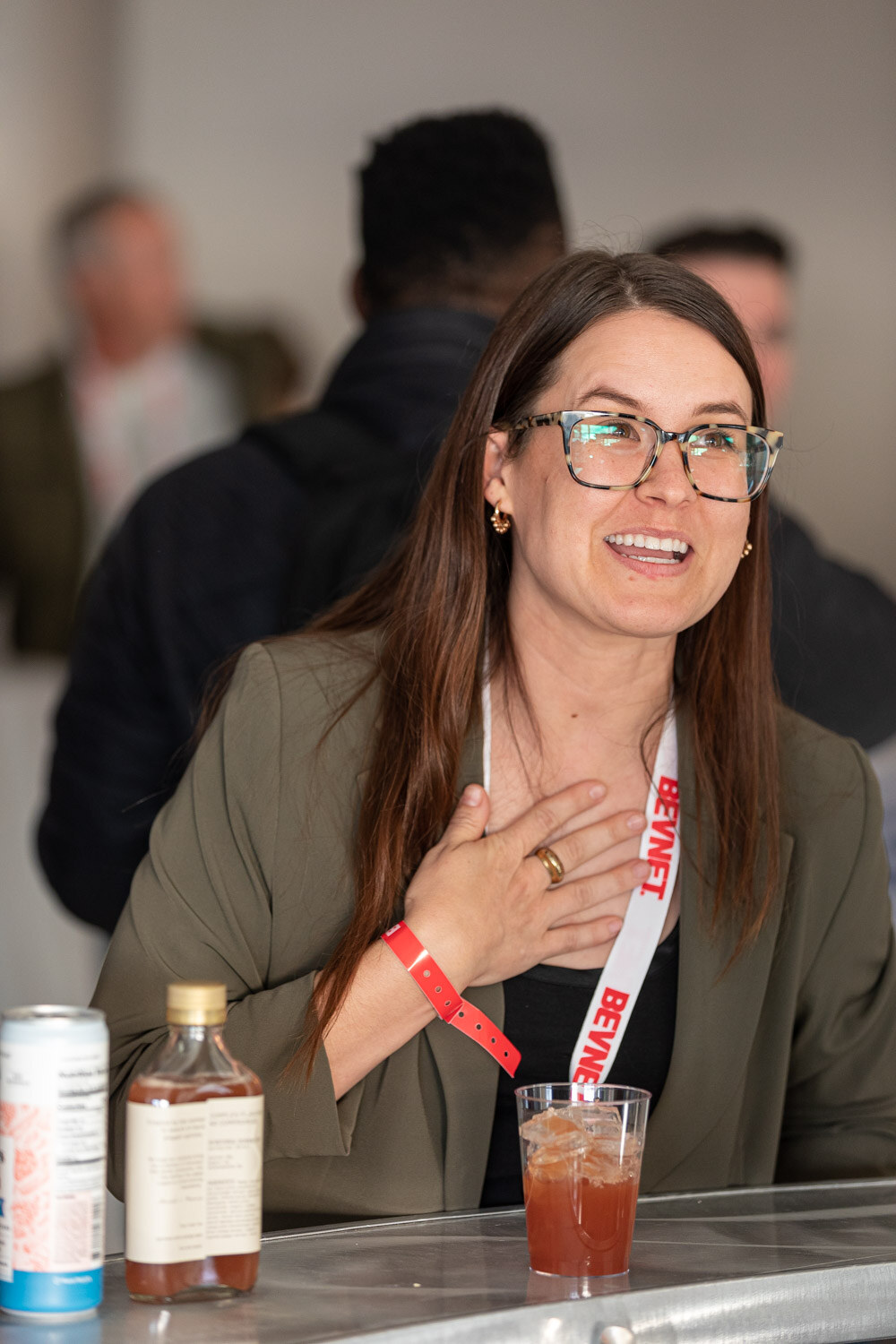 A woman in glasses and an olive jacket with a BevNET lanyard smiles with her hand on her chest, with a bottle and a drink on the counter.