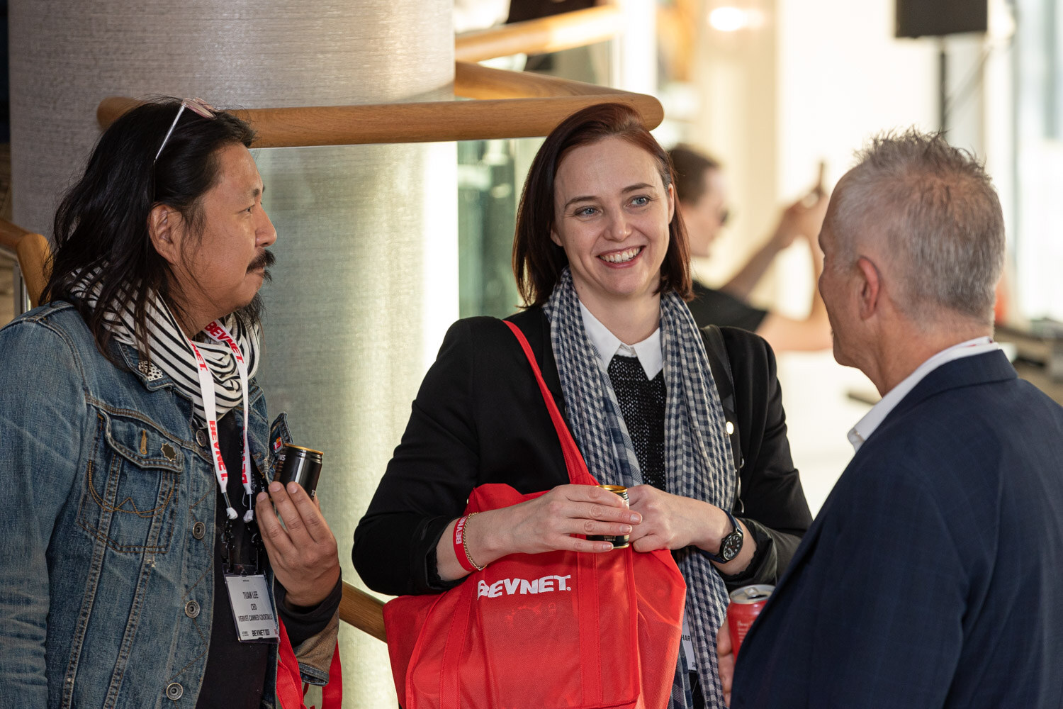 A woman in a black blazer with a red BevNET tote bag talks with a man in a denim jacket and a man in a navy blazer, holding cans.
