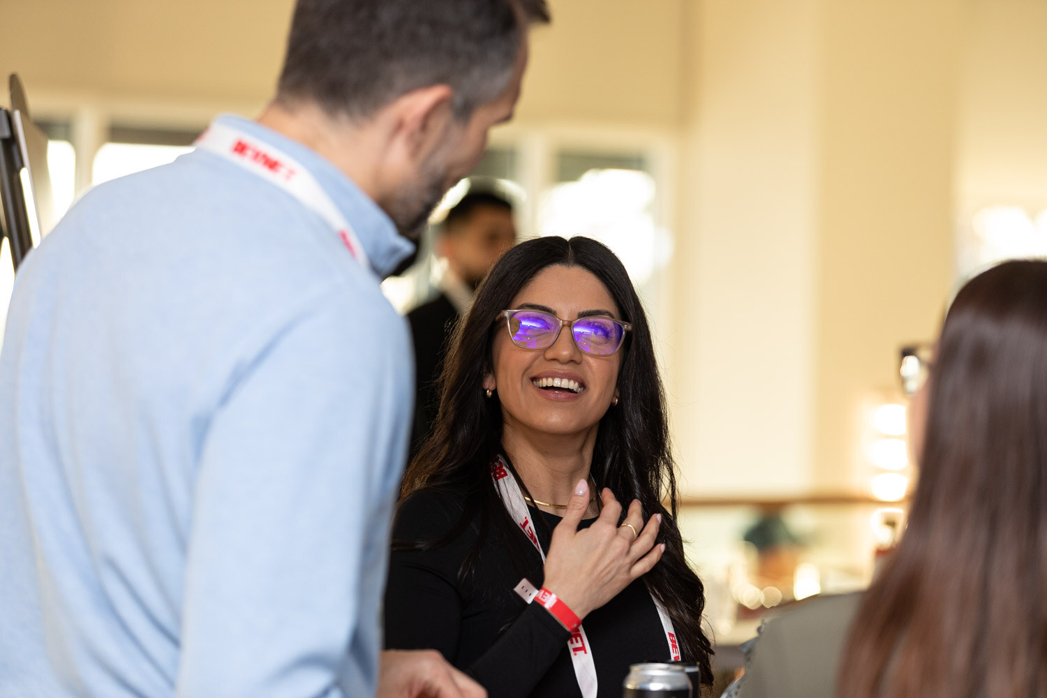 A woman in purple-framed glasses and a black top with a BevNET lanyard laughs with her hand on her chest as a man in a light blue shirt talks.